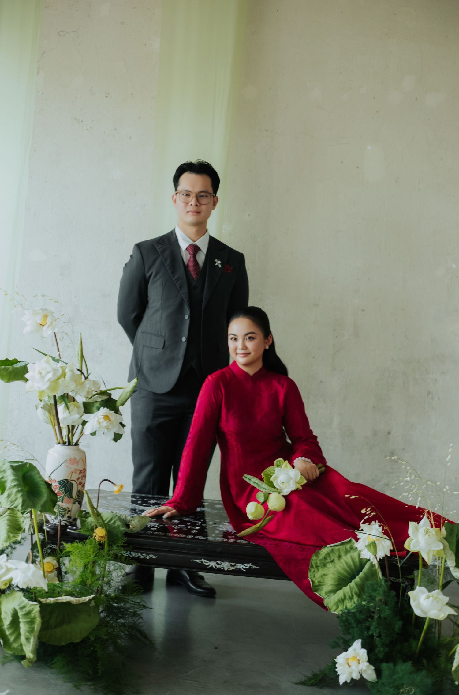 A couple in formal attire pose with lotus flowers, the woman in a red ao dai seated at a traditional instrument.