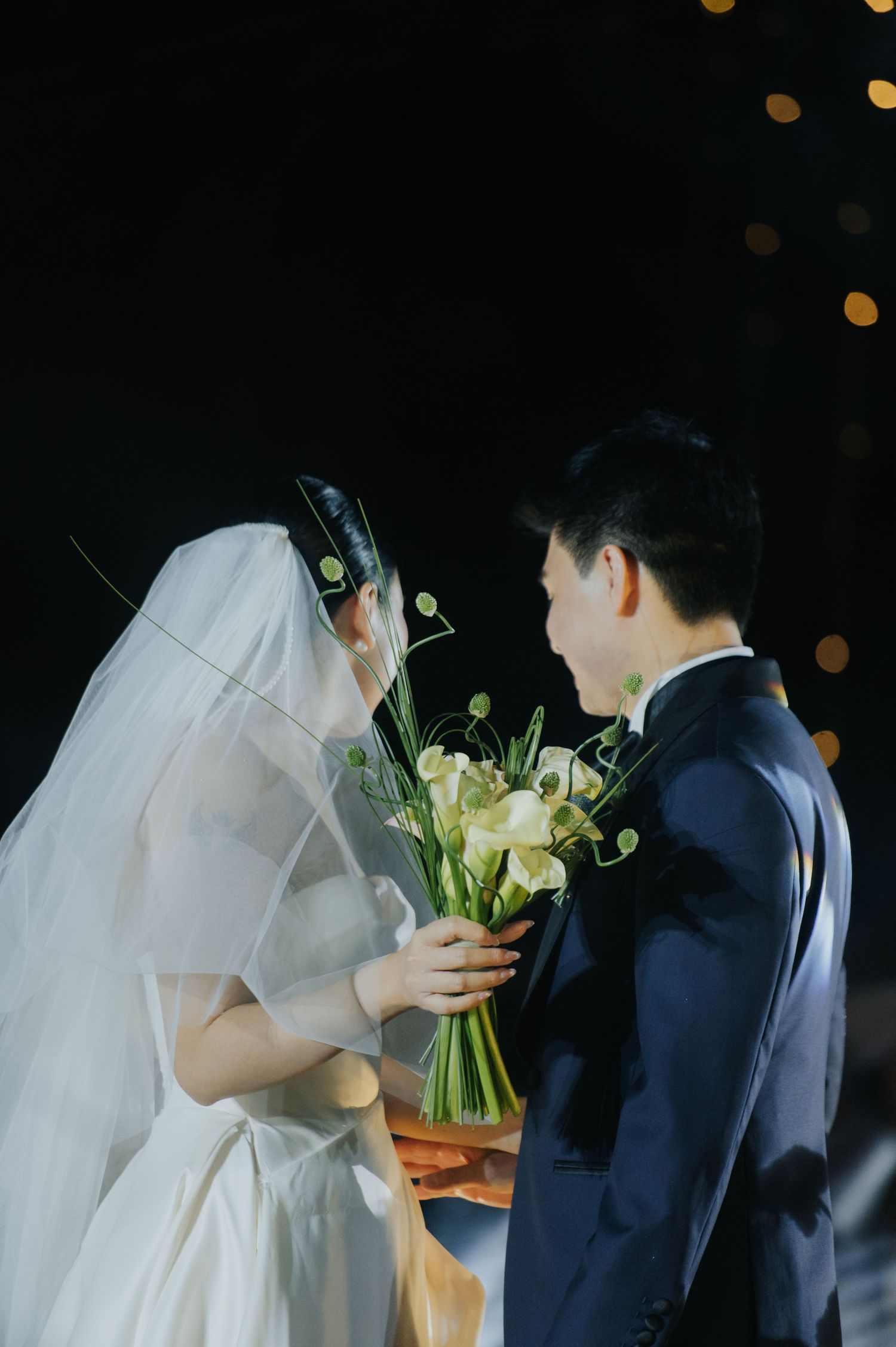 Bride in white gown and veil holds elegant calla lily bouquet with groom in navy suit during wedding ceremony.