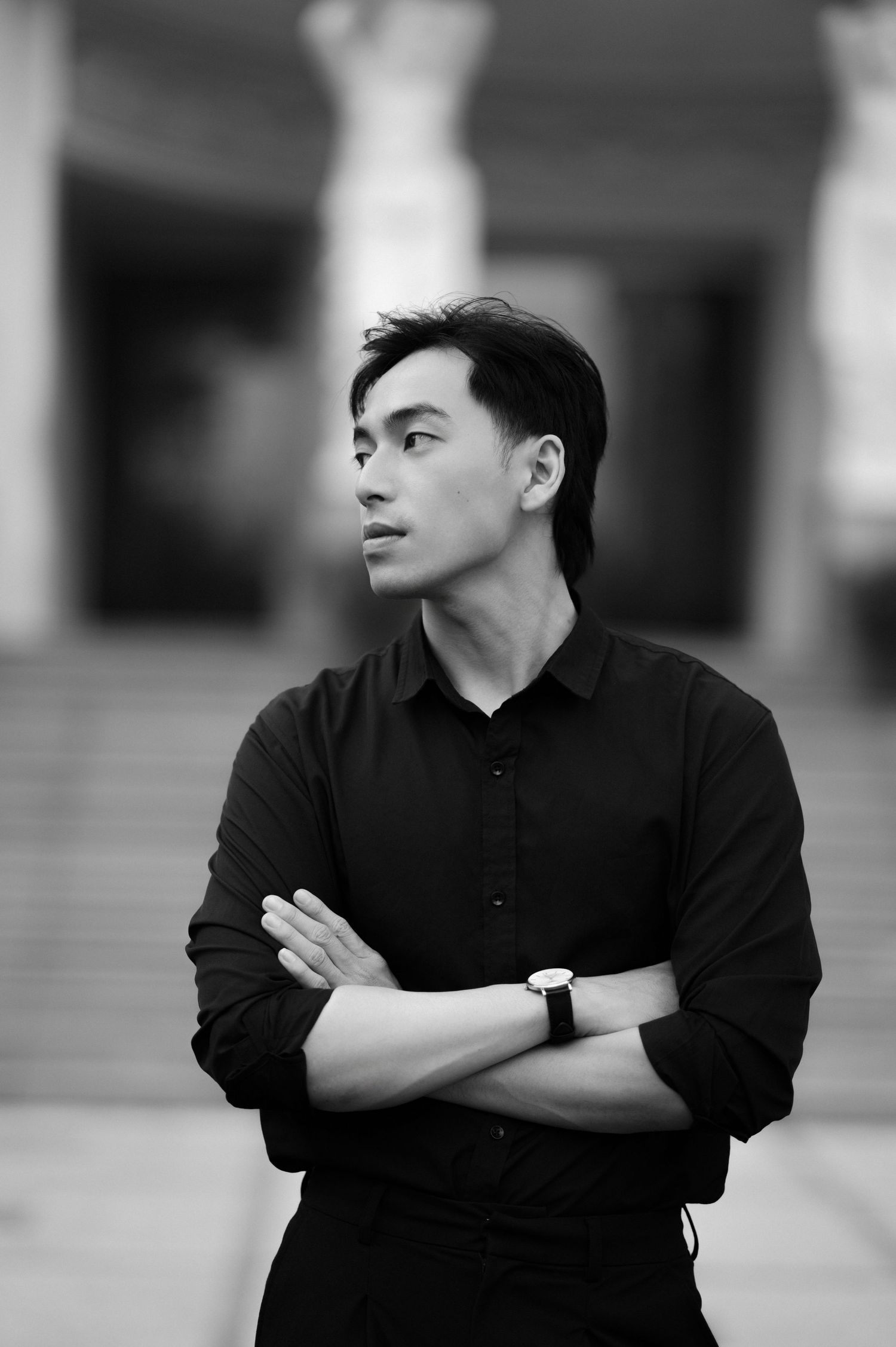 Black and white portrait of a young Asian man in a black shirt with arms crossed, wearing a watch, looking away thoughtfully.