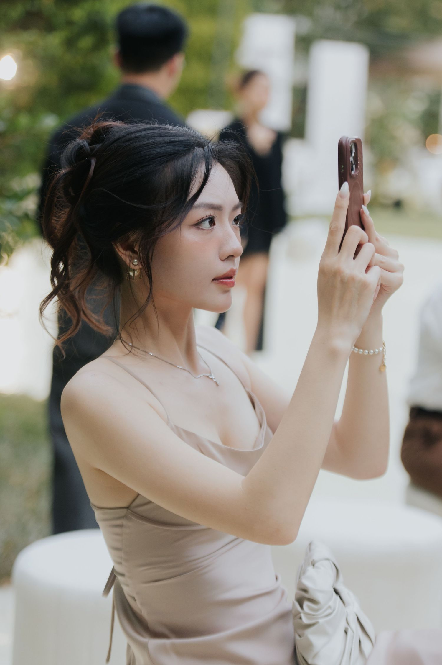Young Asian woman in elegant beige slip dress taking a photo with her smartphone at an outdoor event.