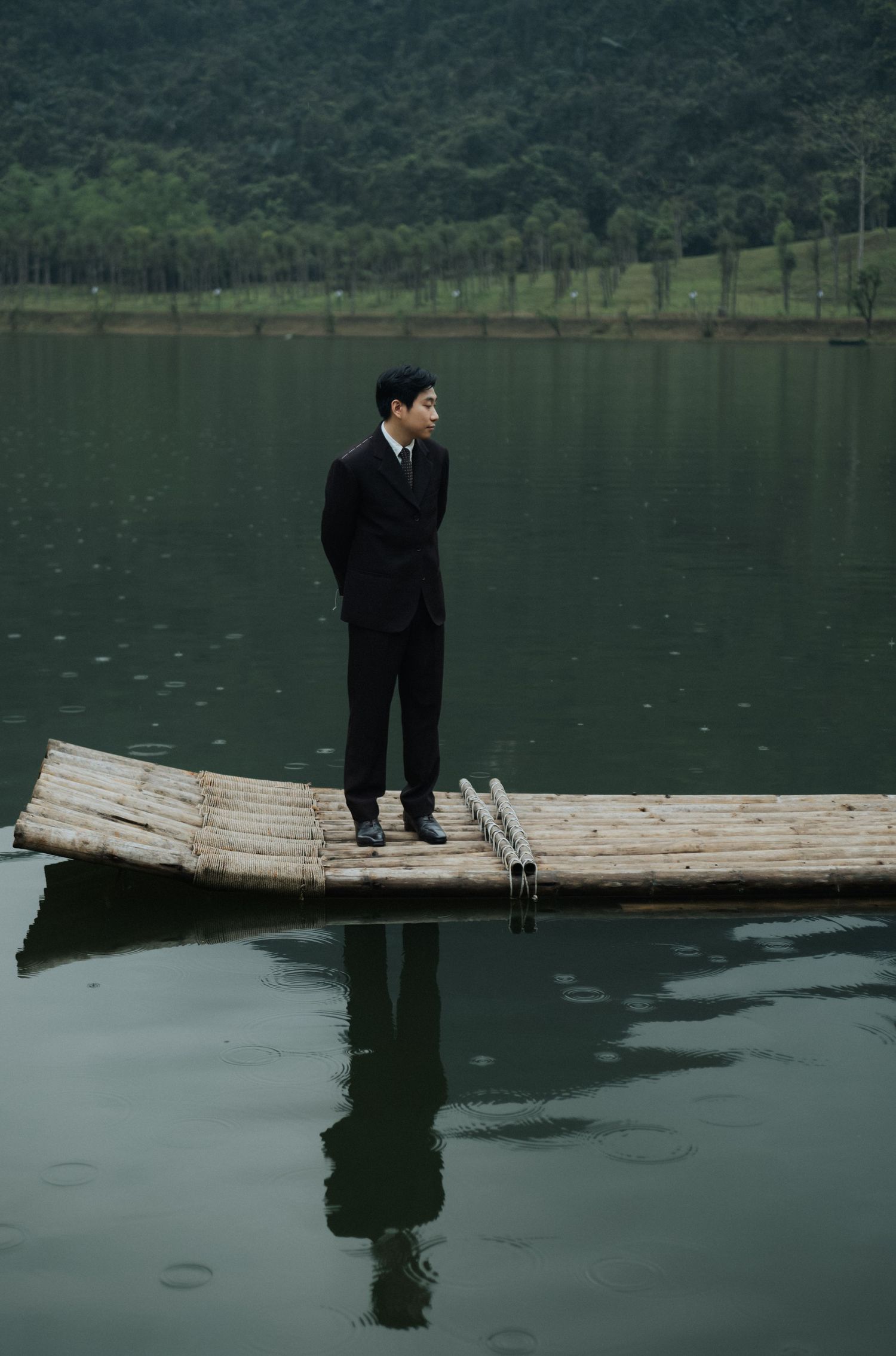 A man in a black suit stands on a bamboo raft on a calm lake with green hills in the background.