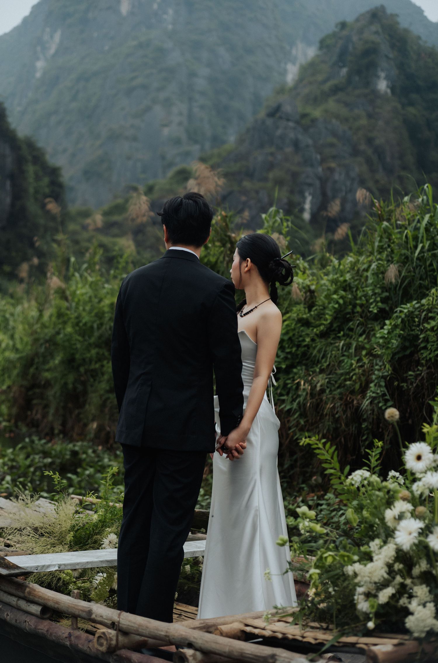 A couple in wedding attire holds hands on a bamboo boat, surrounded by lush greenery and karst mountains in Vietnam.