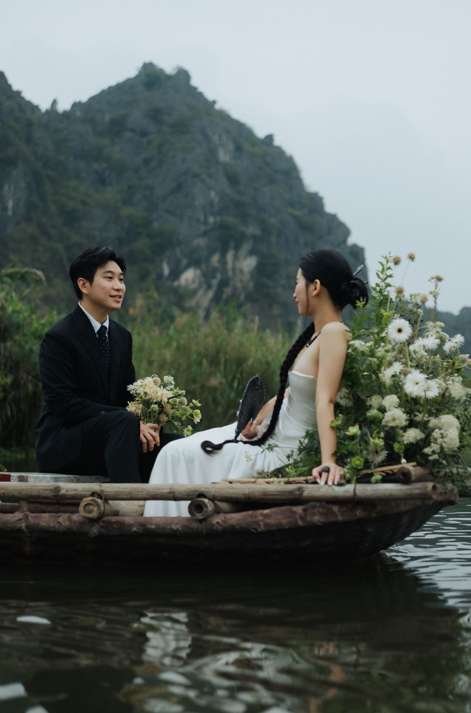 A couple in wedding attire sits on a wooden boat surrounded by white flowers, with misty karst mountains in the background.