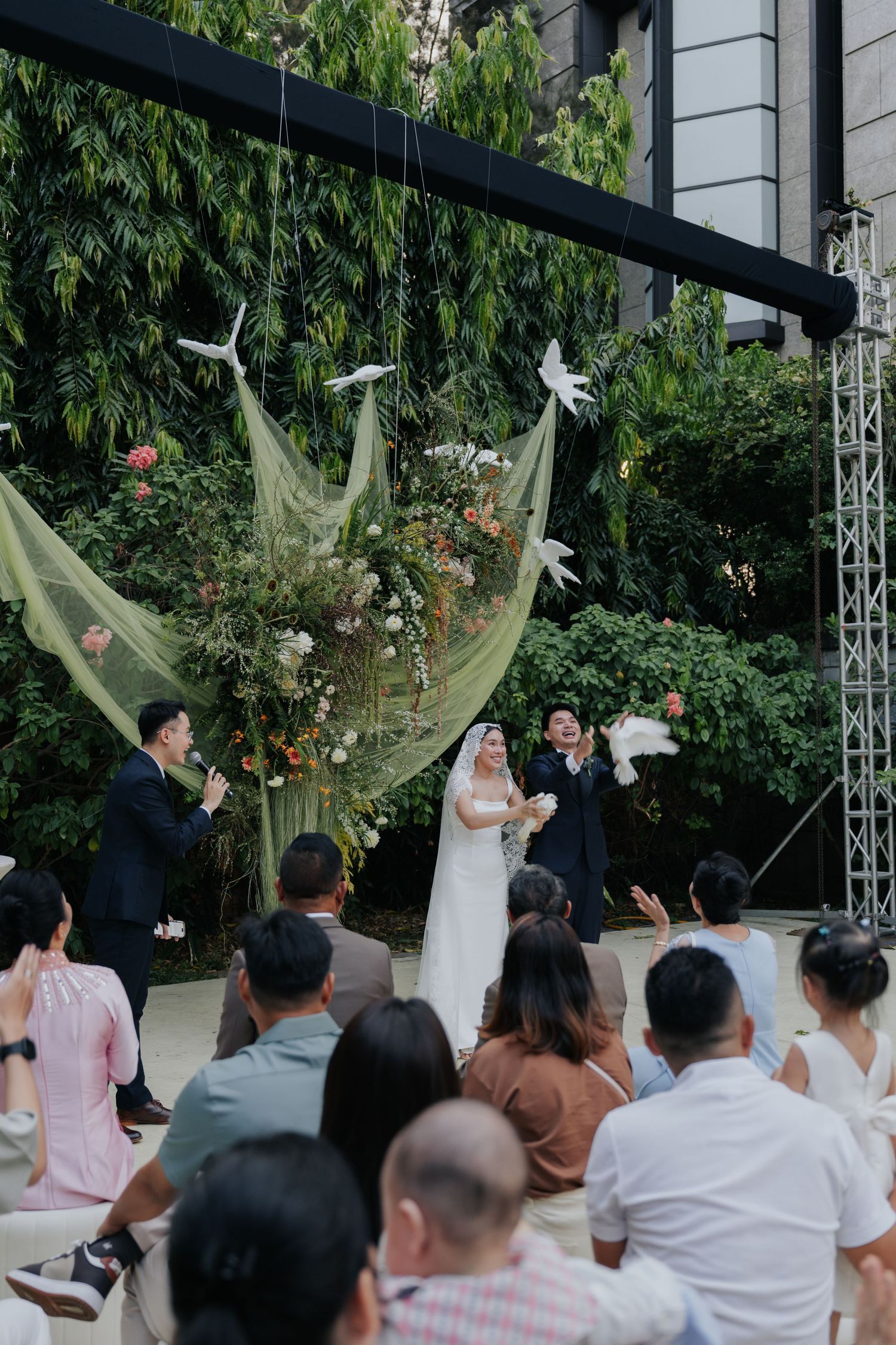 Outdoor wedding ceremony with couple releasing white doves, green floral backdrop, and seated guests in attendance.