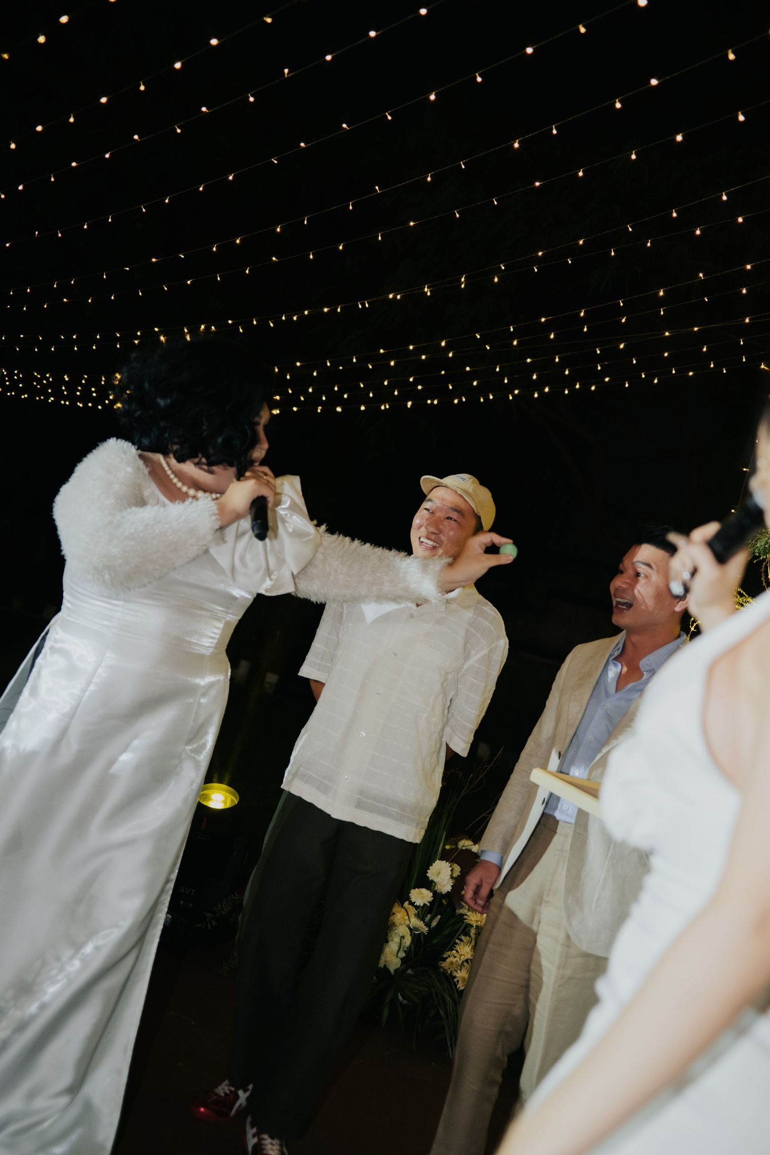 Bride in white gown holds microphone at wedding reception under twinkling star lights with guests laughing nearby.