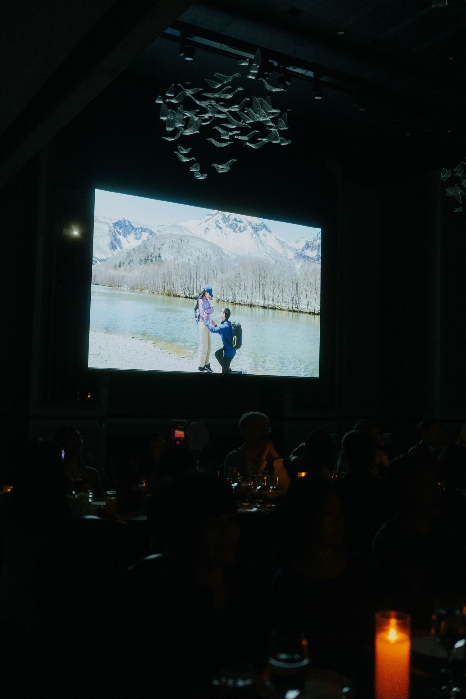 A large projection screen displays a couple near a mountain lake during an elegant candlelit event in a darkened venue.