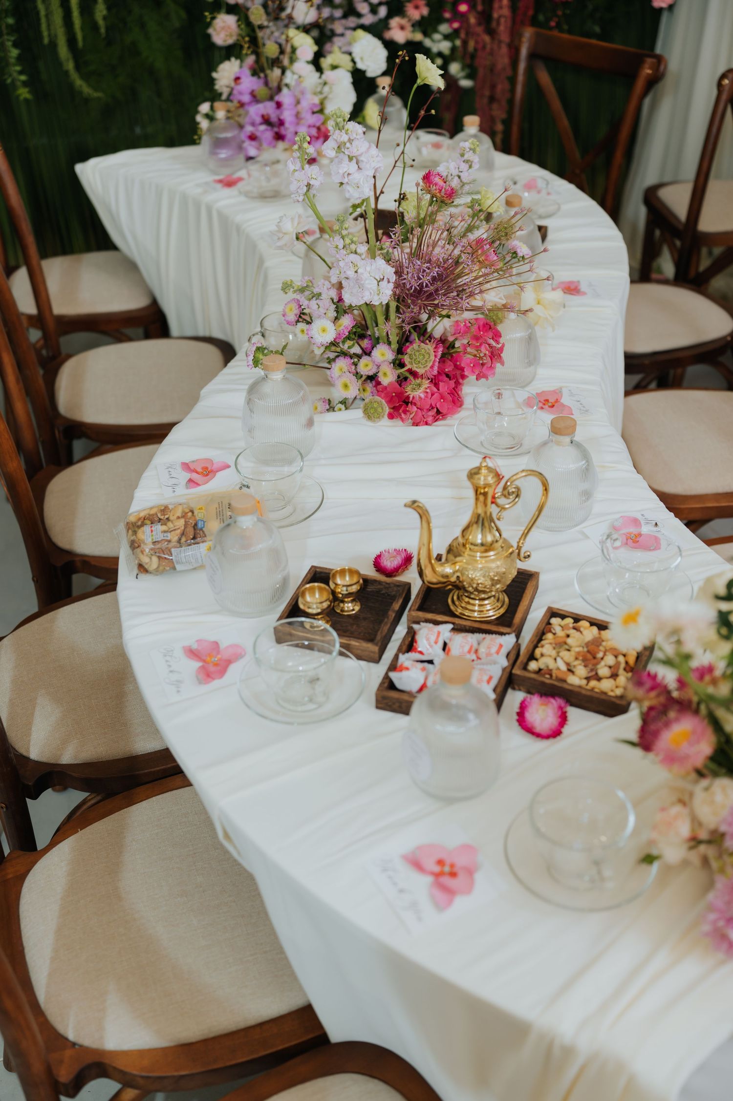 Elegant wedding table with white linen, gold teapot, pink floral arrangements, and wooden chairs for a romantic event.