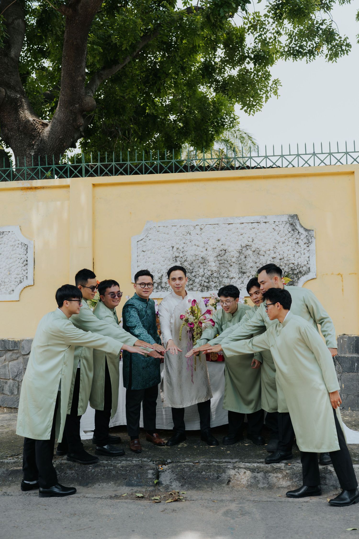 Groomsmen in mint green ao dai gather in a circle around the groom holding a floral bouquet outdoors.