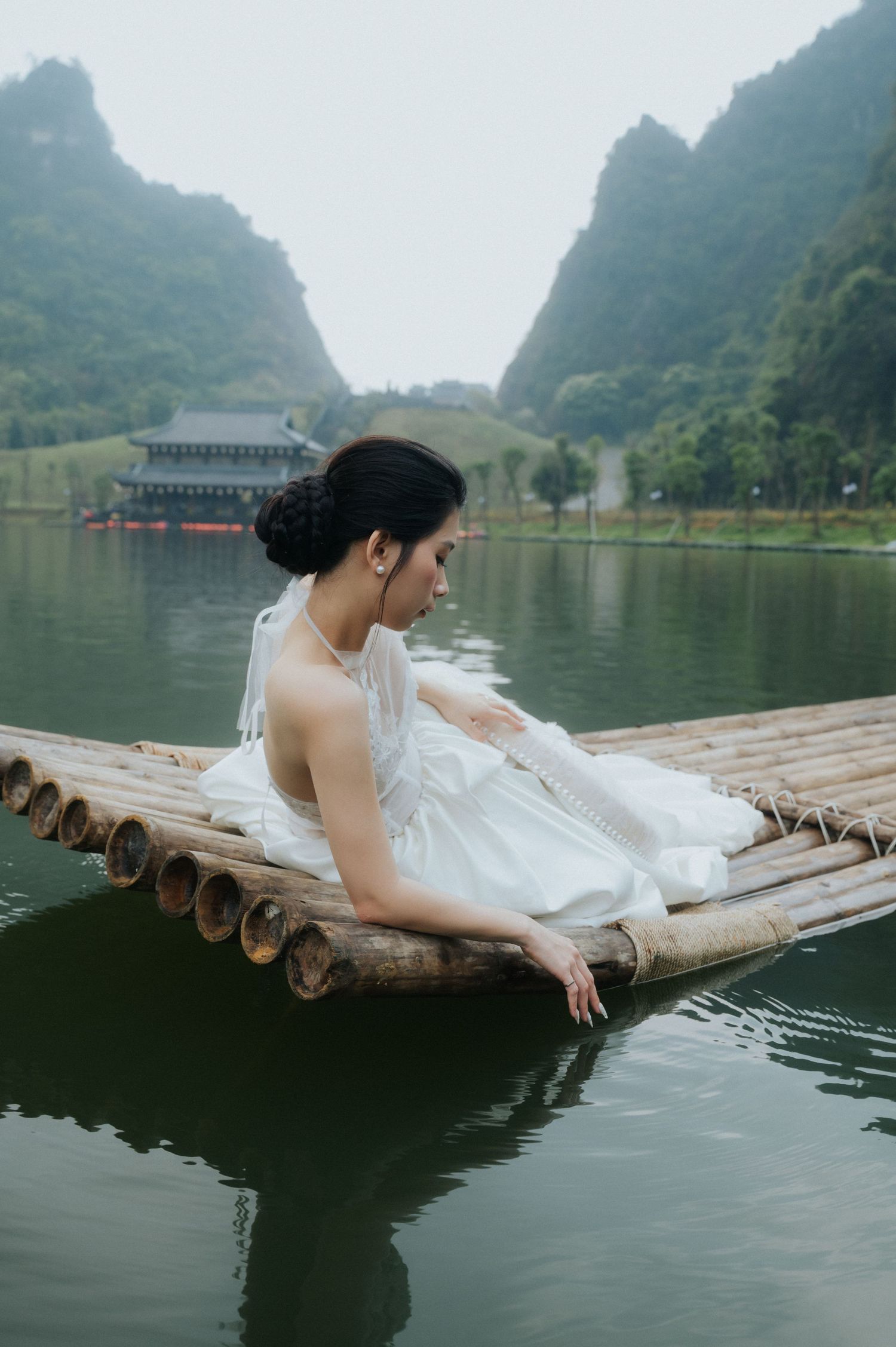 A woman in a white dress sits gracefully on a bamboo raft over calm water, with misty mountains in the background.