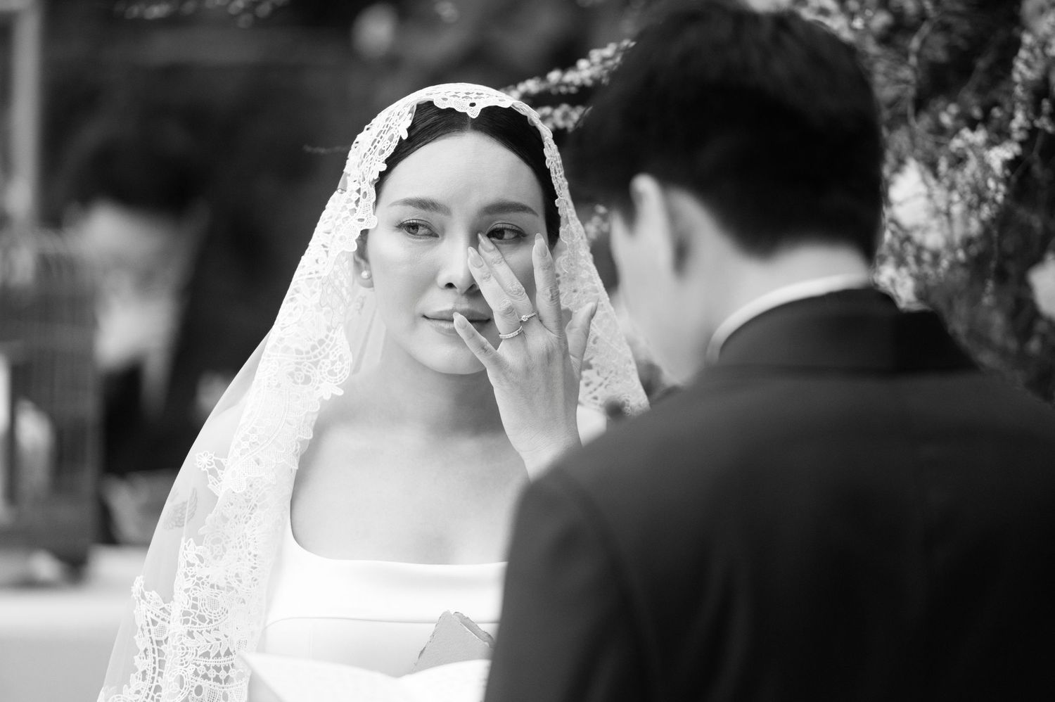 Black and white wedding photo of a bride in veil wiping tears while facing groom in suit during emotional ceremony.