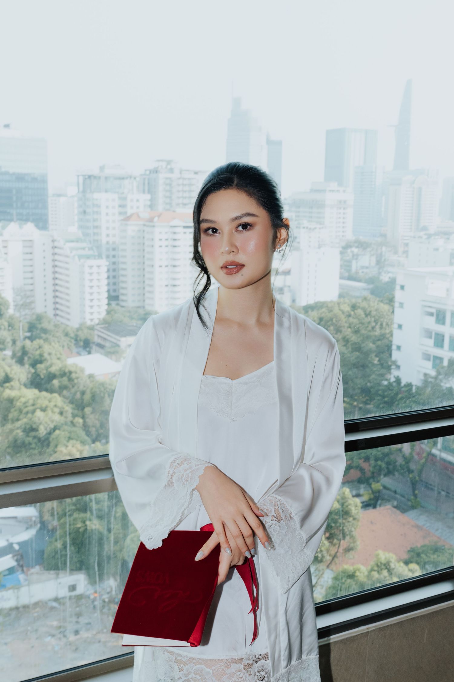 Young woman in white robe holding red clutch, standing by window with city skyline backdrop.