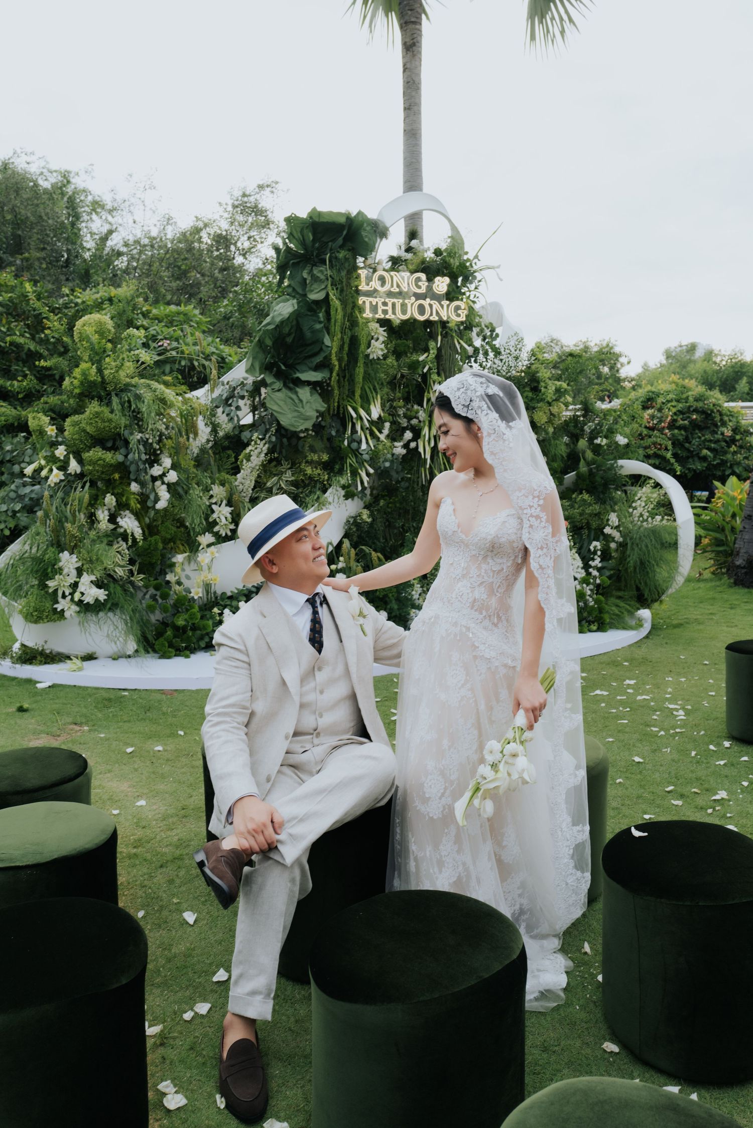 A couple in wedding attire poses outdoors, groom in white suit and hat, bride in lace gown holding bouquet, green floral backdrop.