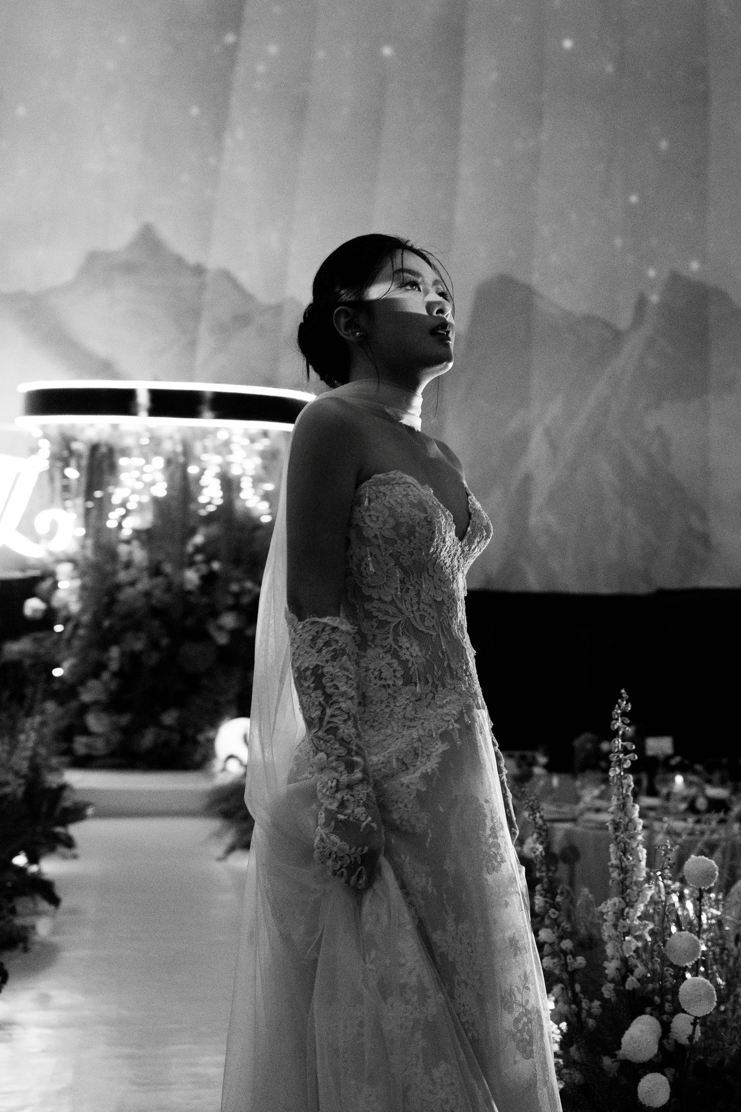 Black and white photo of a bride in a lace wedding gown gazing upward at a decorated wedding venue.