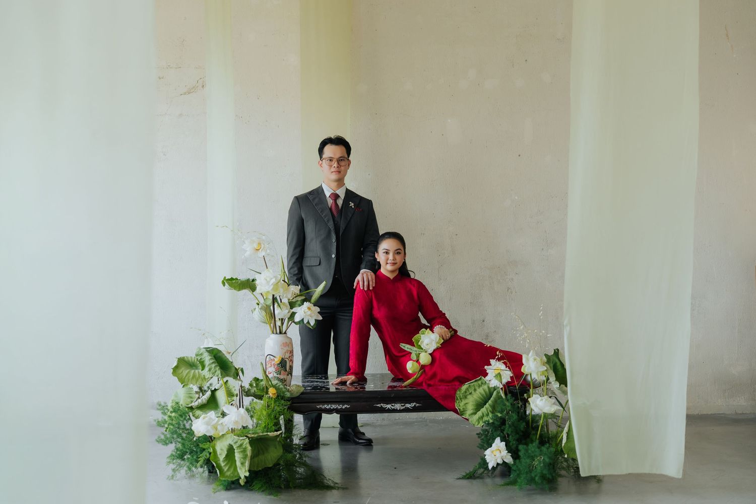 Elegant couple in formal attire posing with white floral arrangements in a minimalist studio setting.