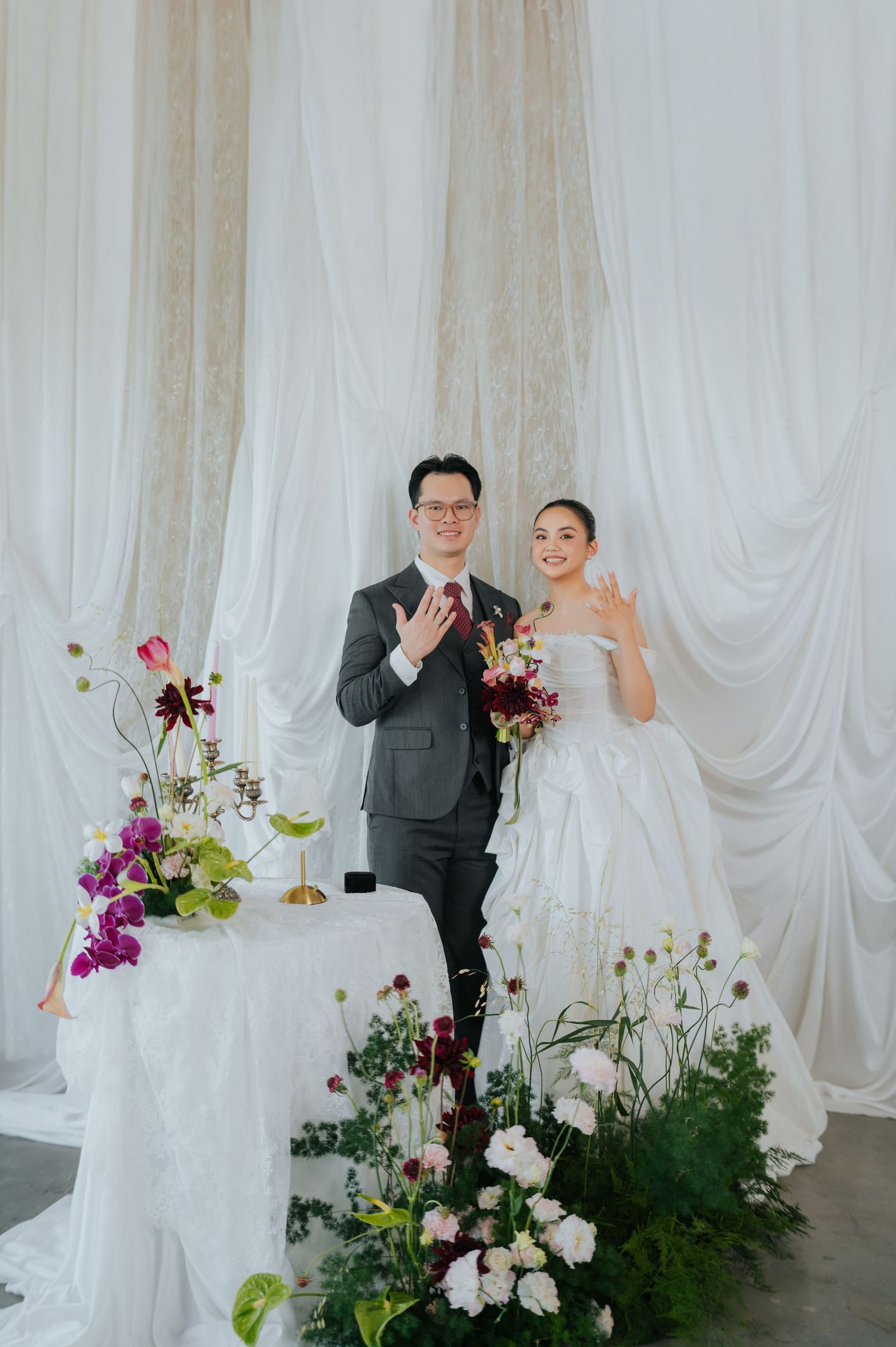 Smiling bride and groom show off wedding rings at elegant floral altar with white drape backdrop.