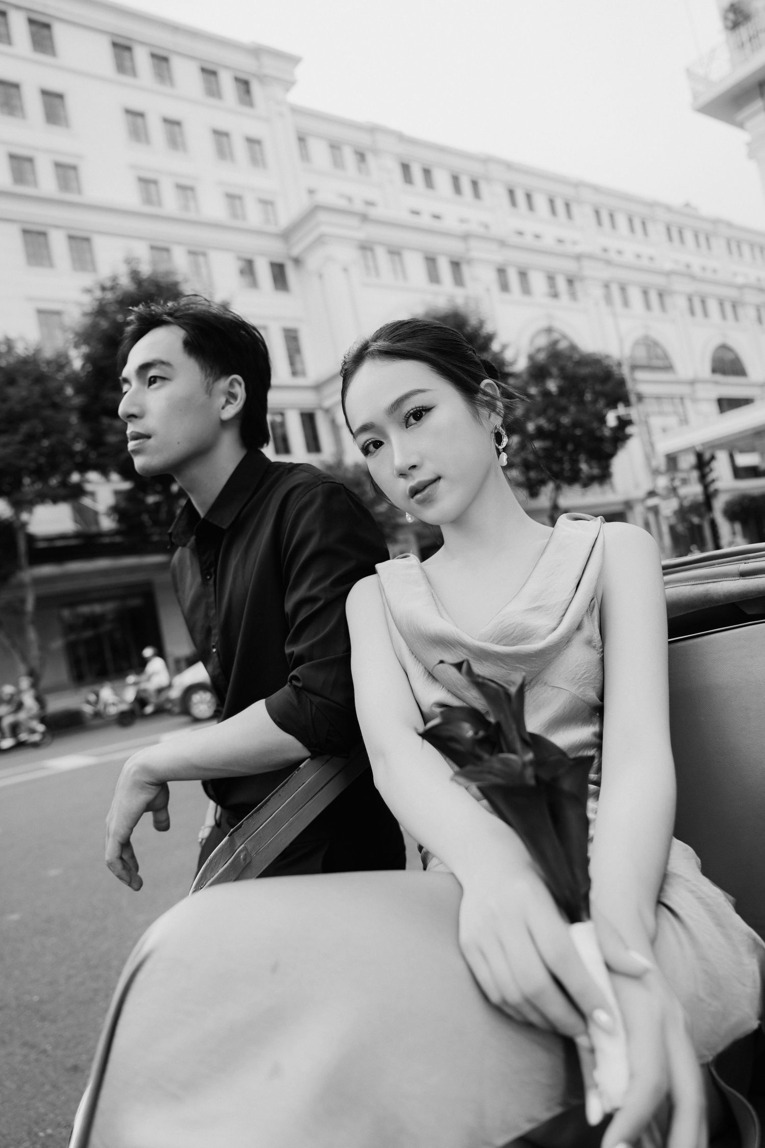 Black and white photo of a young Asian couple sitting on a vintage car on a city street.