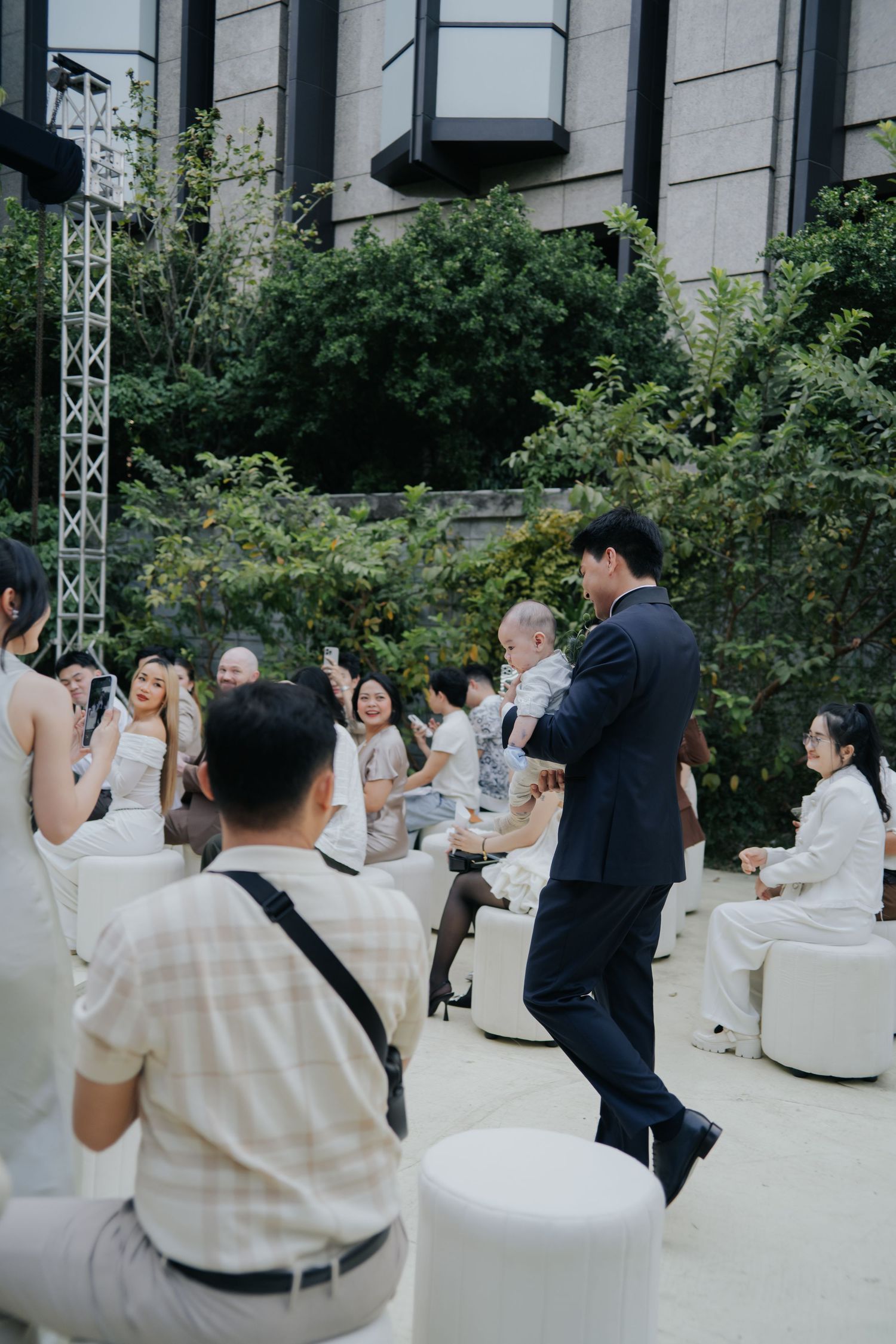 A man in a dark suit dances at an outdoor event while guests in white attire sit and smile around him.