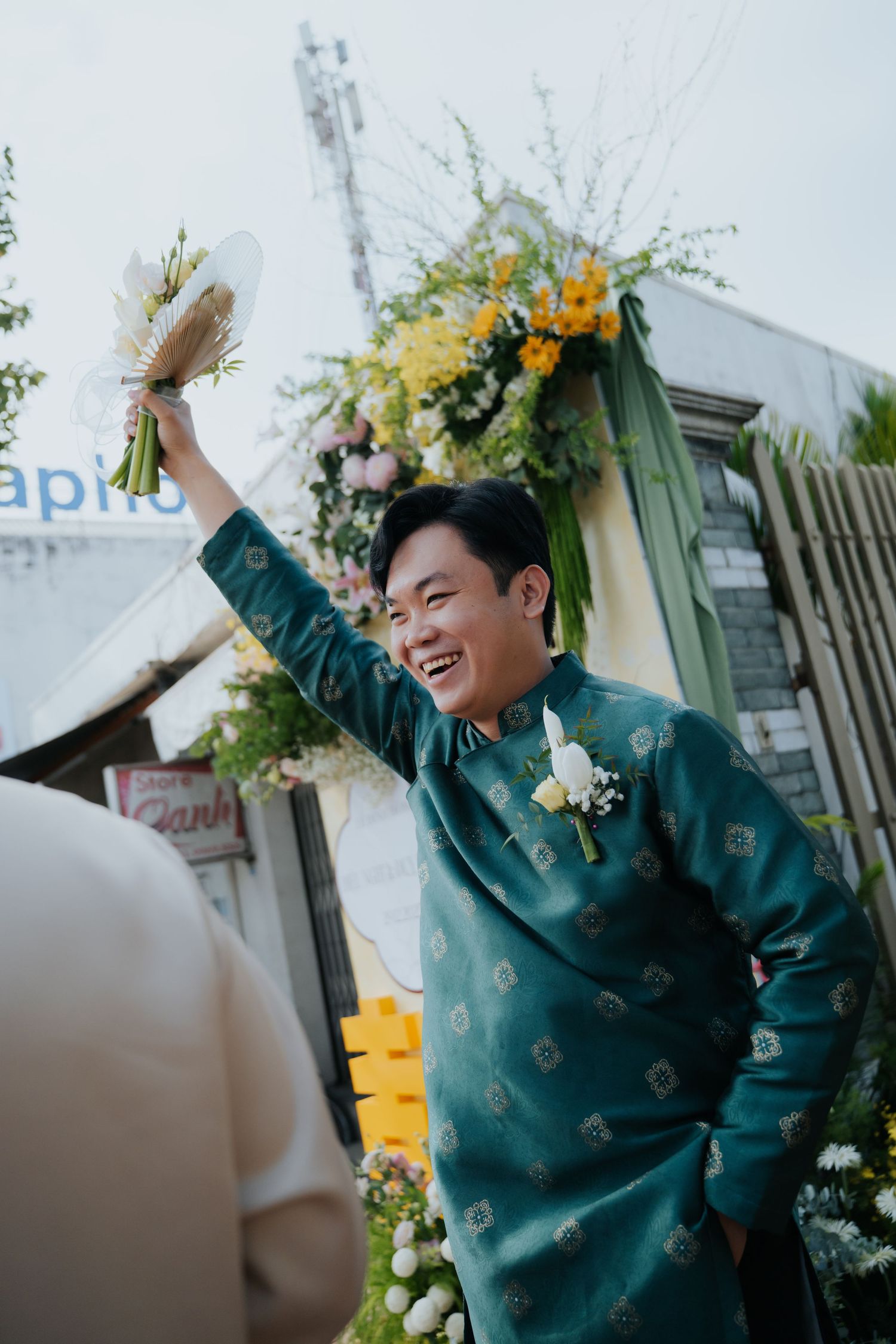 Joyful groom in teal ao dai raises bouquet at Vietnamese wedding ceremony with floral decorations.