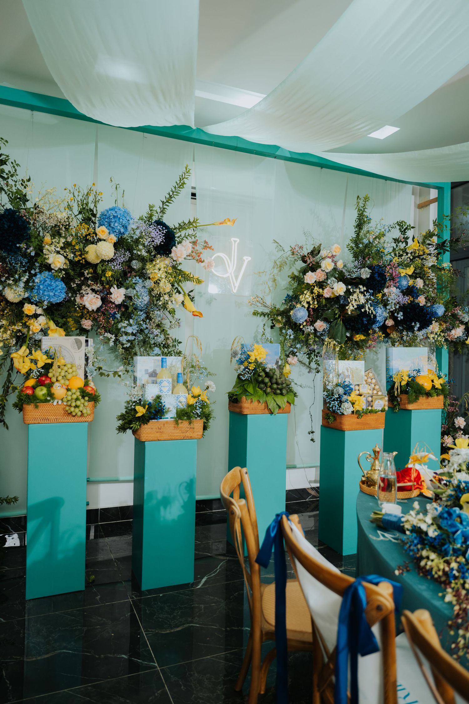 Elegant wedding decor with teal pedestals, blue and yellow floral arrangements, and draped white ceiling in a reception venue.