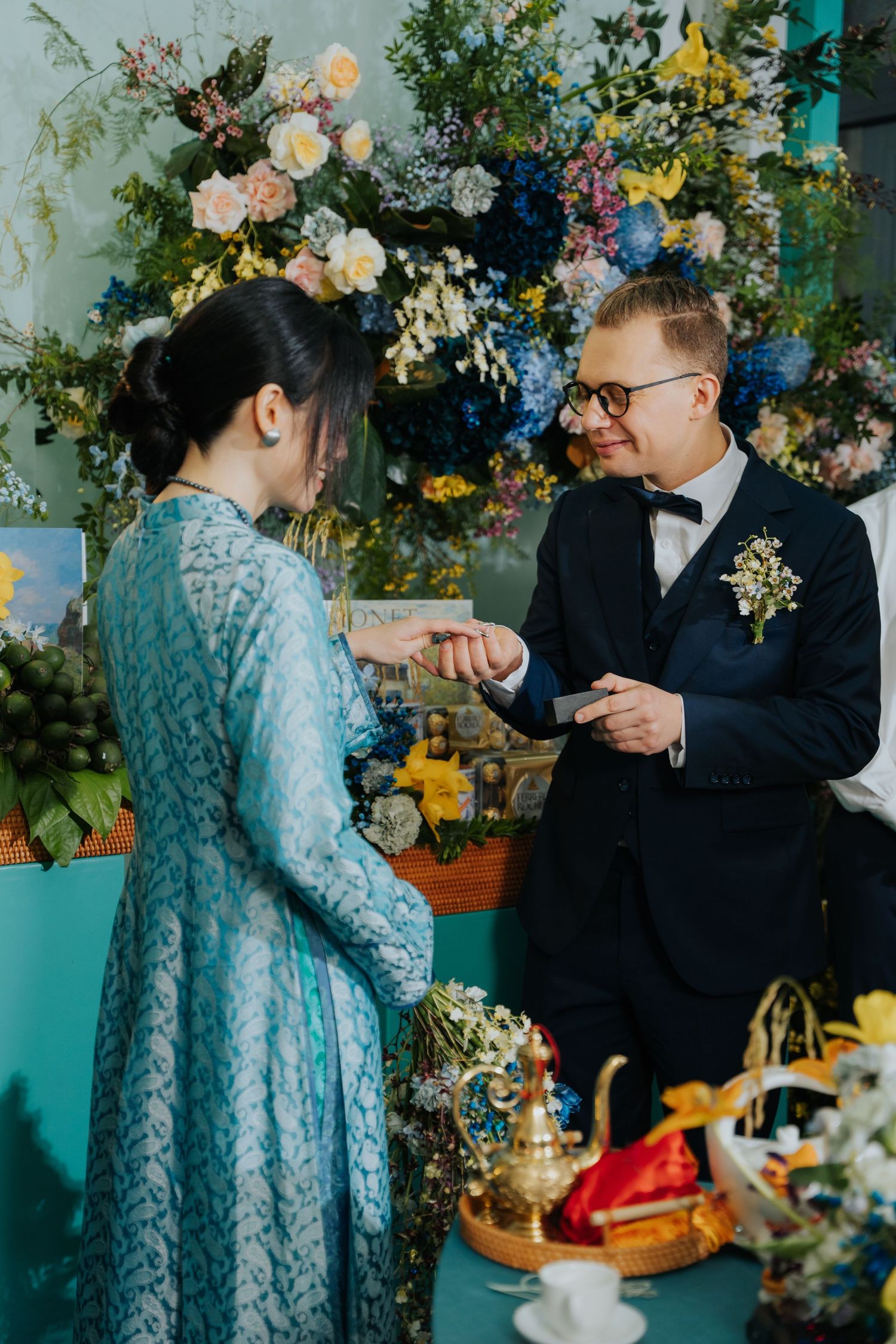 Groom in navy suit placing wedding ring on brides finger at floral altar during traditional ceremony.