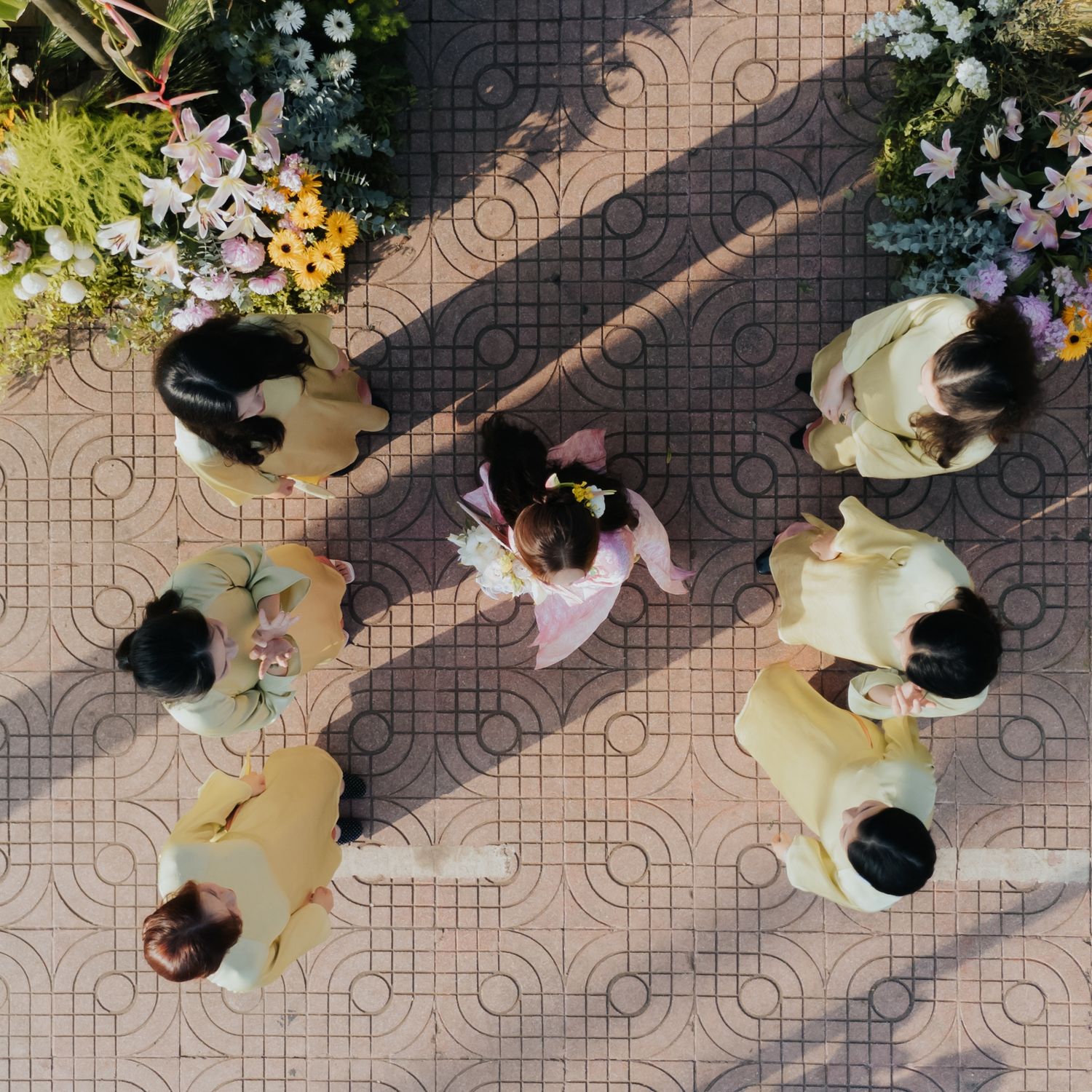 Aerial view of children in yellow outfits gathered in a circle around a girl in pink on a patterned floor.