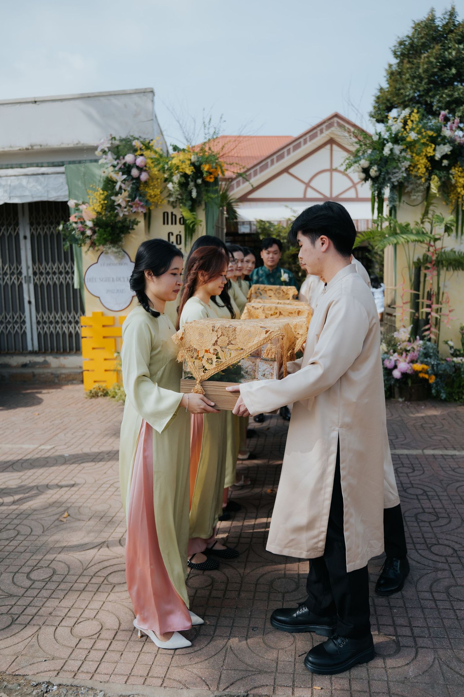 People in traditional Vietnamese ao dai exchanging ornate gifts at an outdoor ceremony with floral decorations.