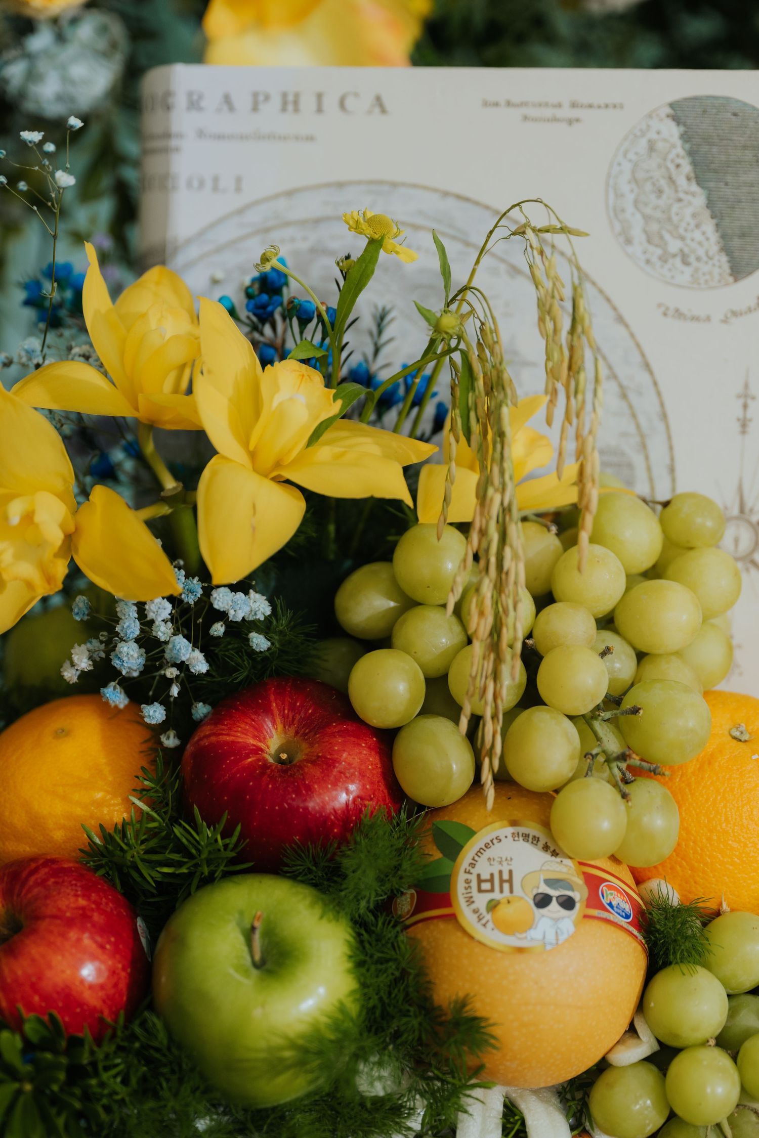Colorful fruit basket with red and green apples, grapes, oranges, and yellow lilies arranged with wheat stalks and blue flowers.