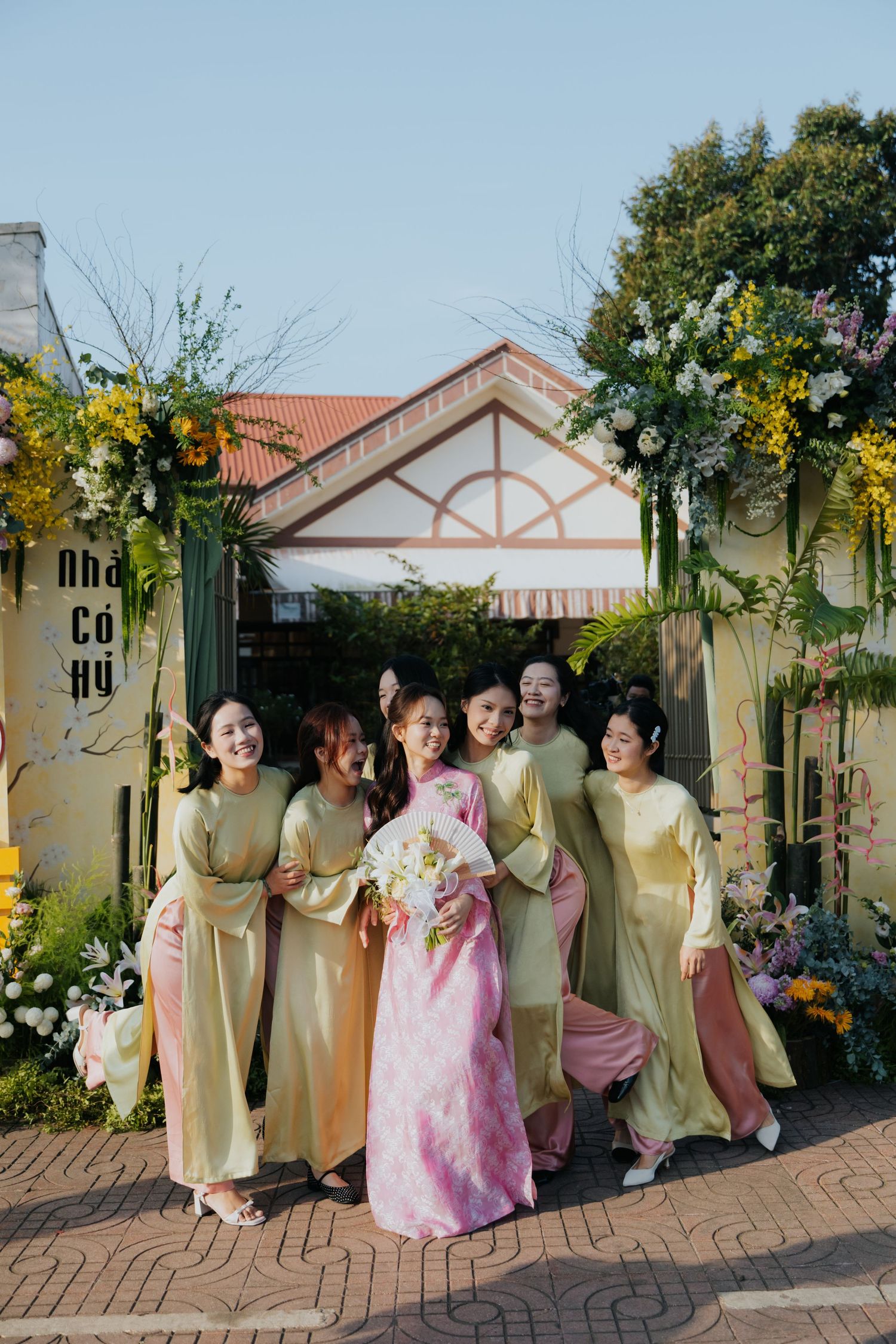 Bride in pink ao dai with bridesmaids in yellow ao dai posing at floral gate in Vietnam.