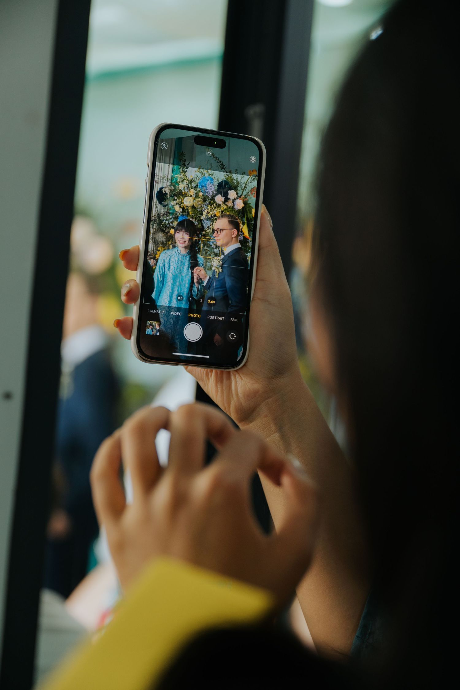 Person photographing a couple posing in front of floral backdrop using an iPhone camera at an event.