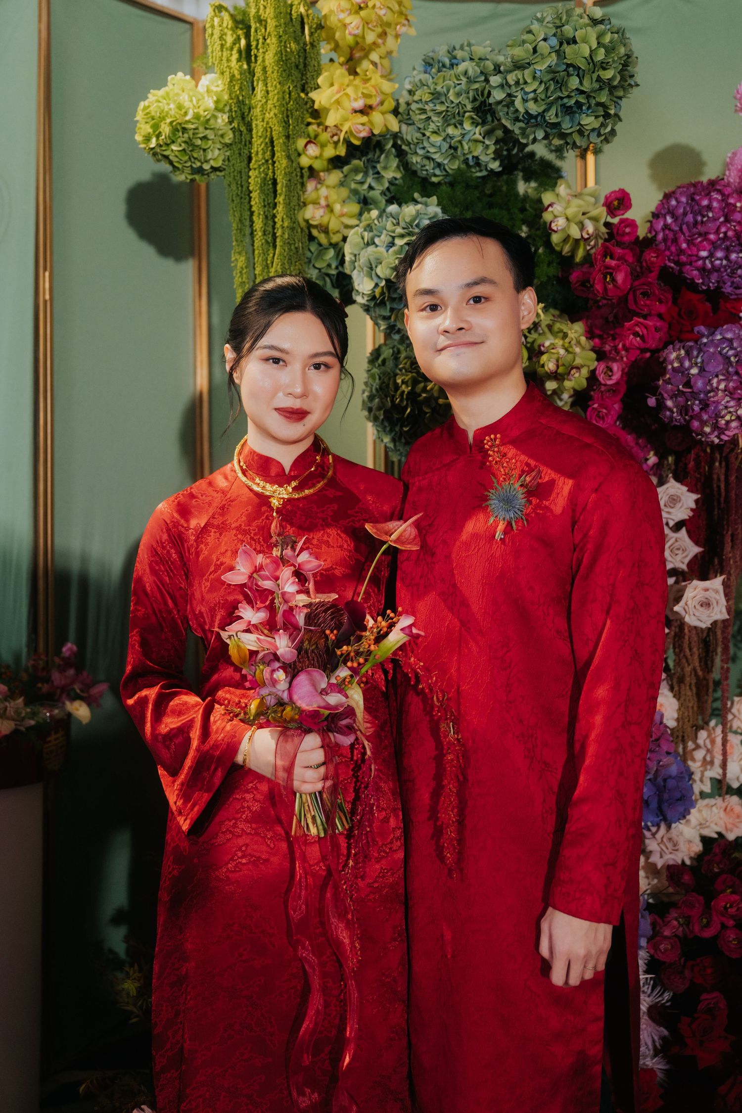 Couple in traditional red Vietnamese ao dai attire posing with floral bouquet at a wedding ceremony.