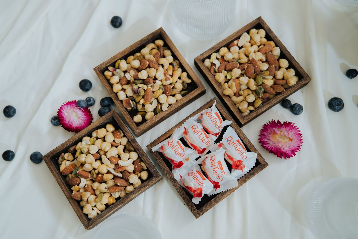 Wooden boxes filled with mixed nuts and dried fruits arranged on white fabric with blueberries and pink flowers.