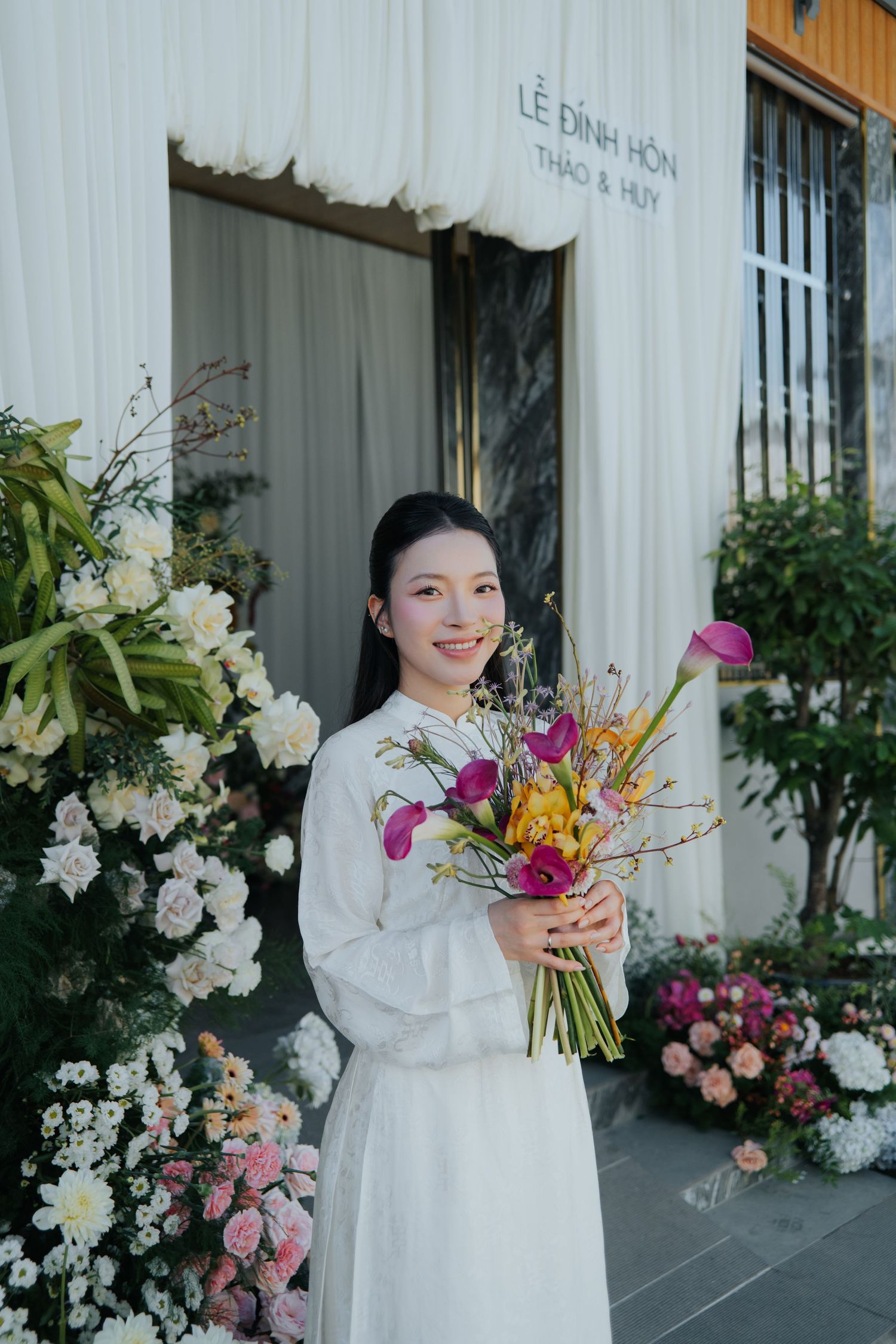 Smiling Vietnamese bride in white ao dai holds colorful bouquet outside floral-decorated wedding venue.