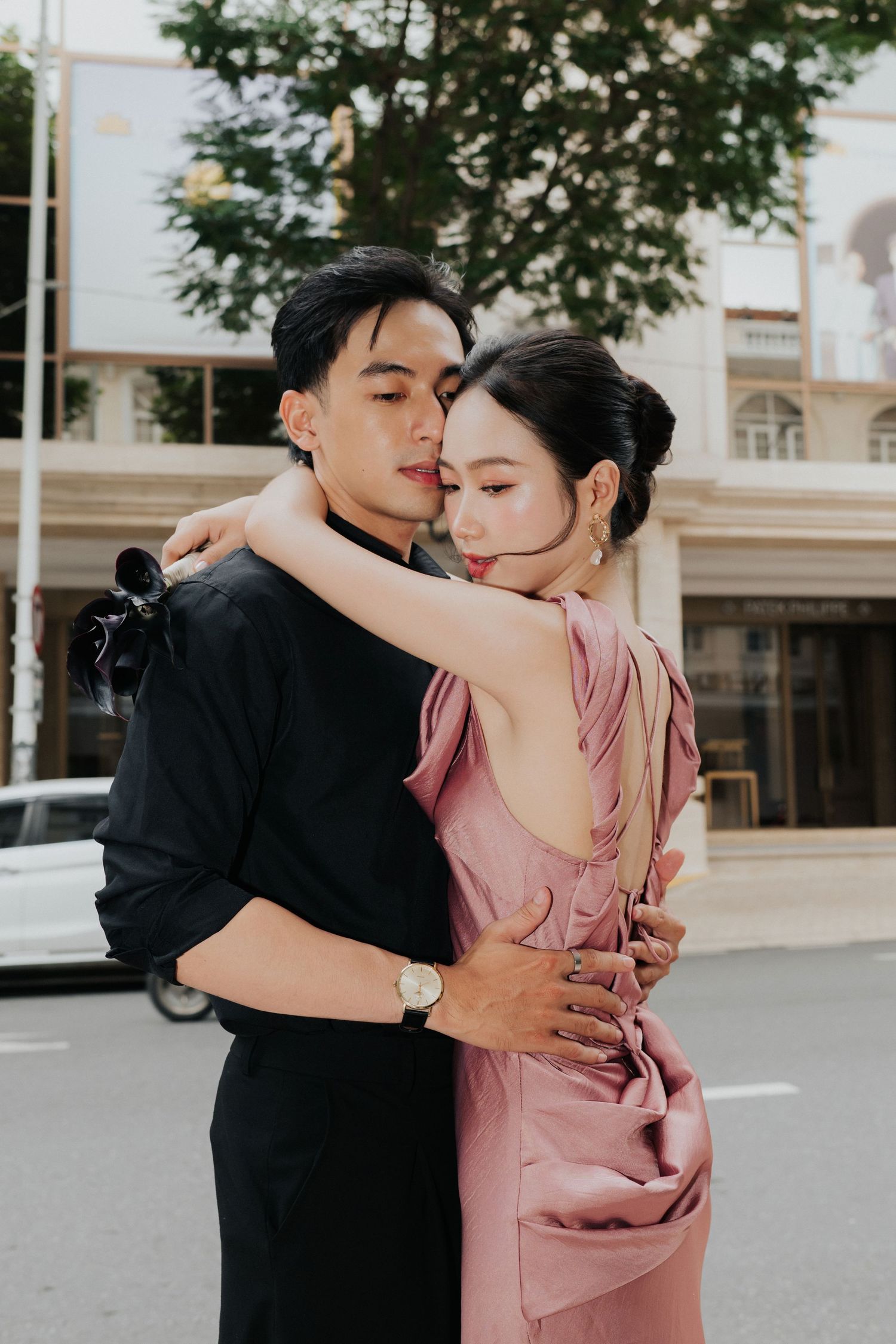A stylish Asian couple embraces on a city street, man in black outfit and woman in elegant pink backless dress.