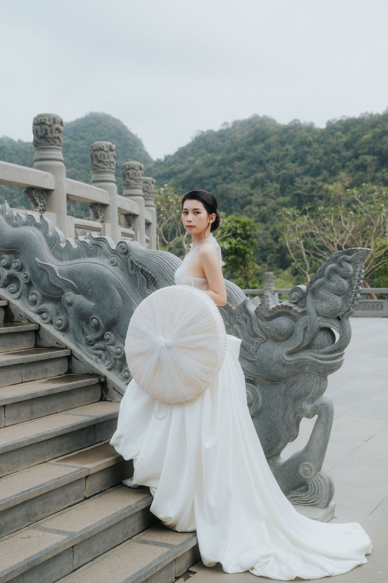 Bride in white gown holds parasol on ornate dragon staircase at Asian temple with misty mountains.