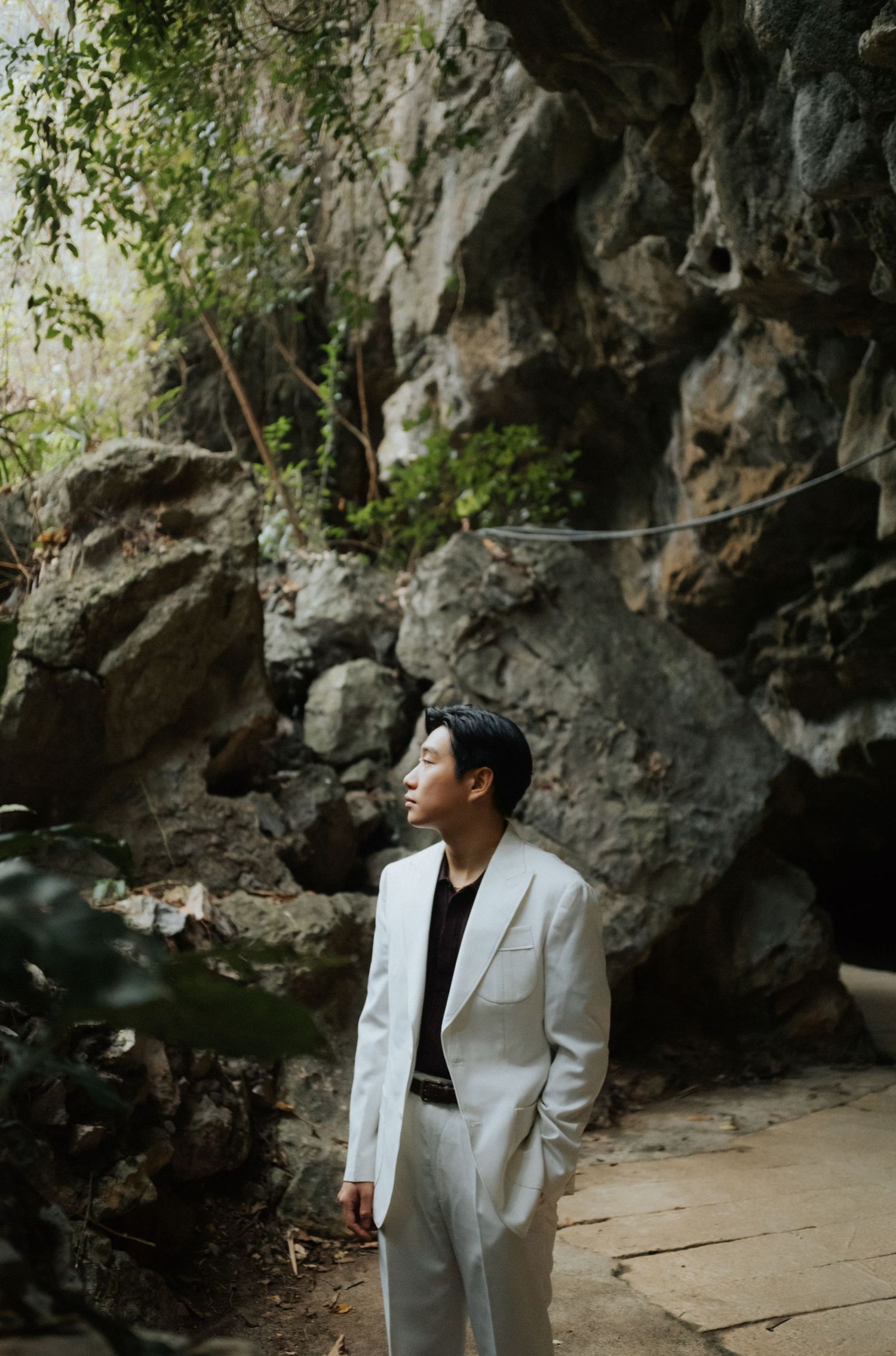 A young man in a white suit stands gazing upward near rocky cliffs and lush greenery outdoors.