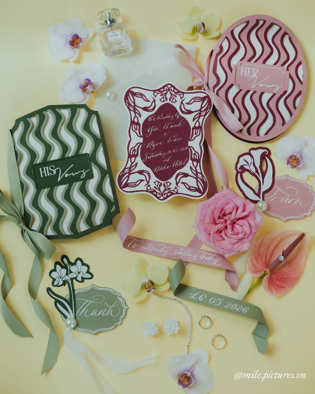 Elegant wedding stationery flat lay with vow books, invitation cards, rings, and floral accents on yellow background.
