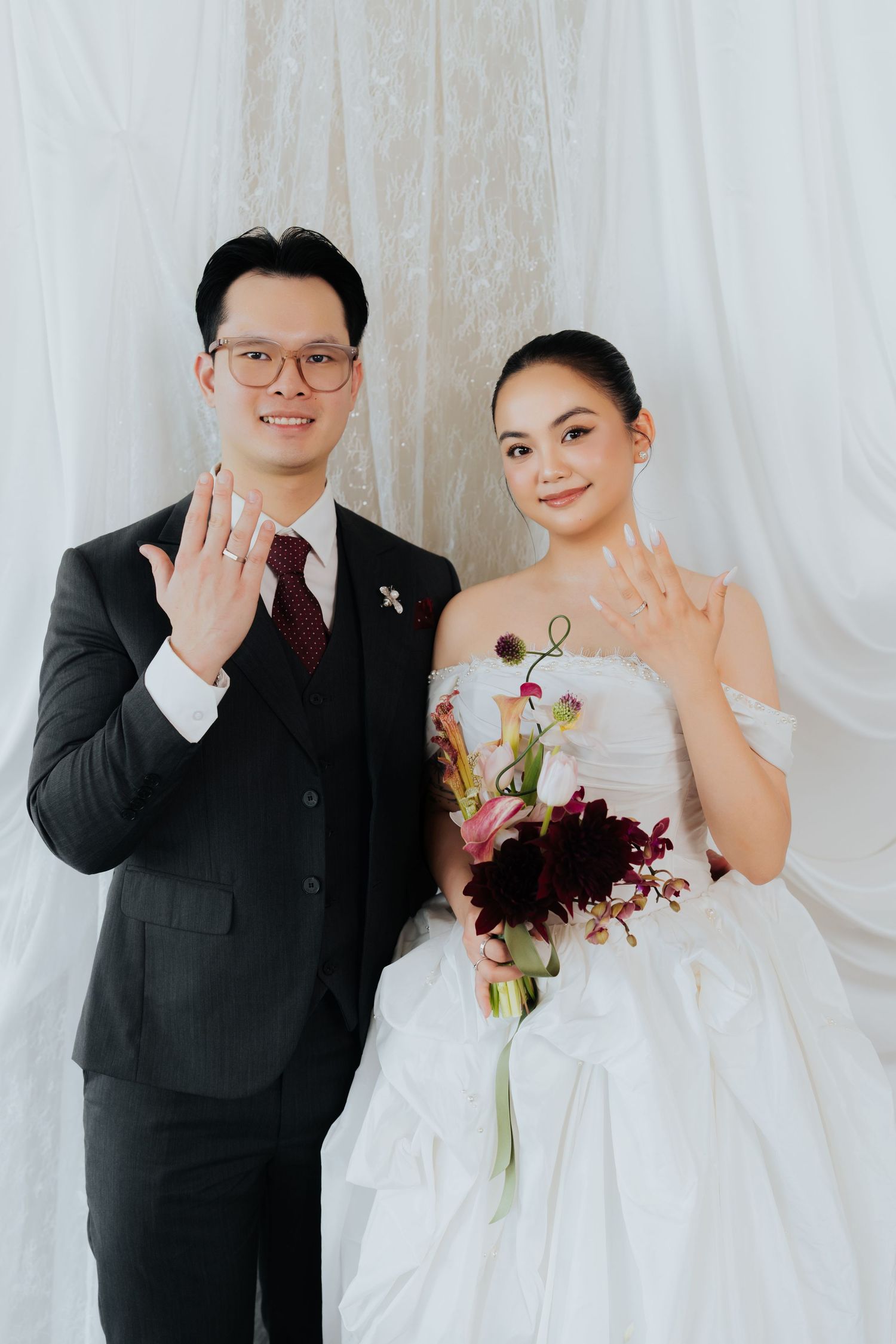 A newlywed couple in wedding attire smiling and showing their rings, bride holding a bouquet.