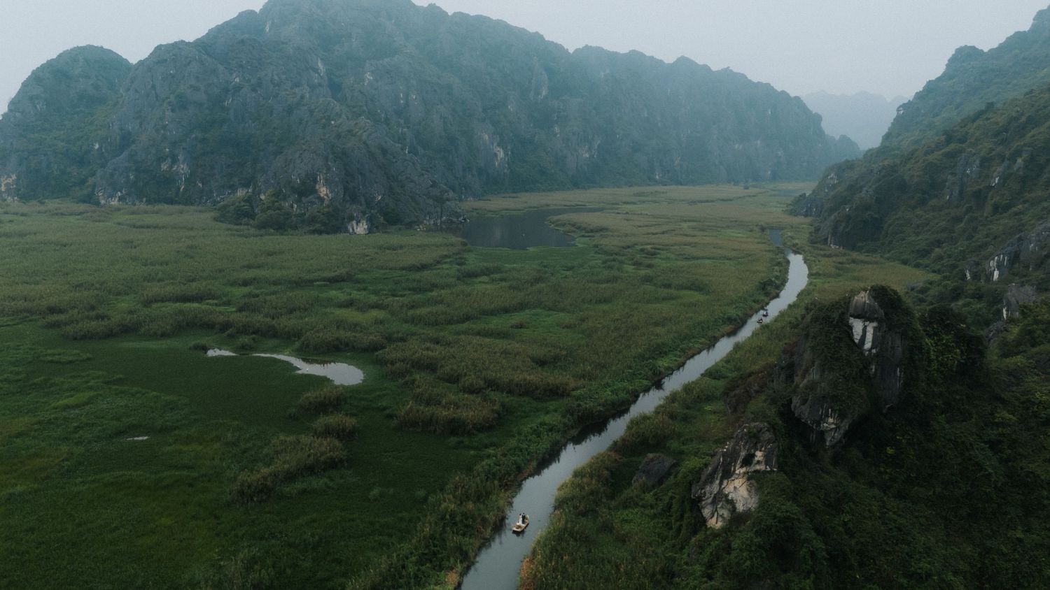 Aerial view of a winding river through lush green valley surrounded by misty limestone karst mountains in Vietnam.