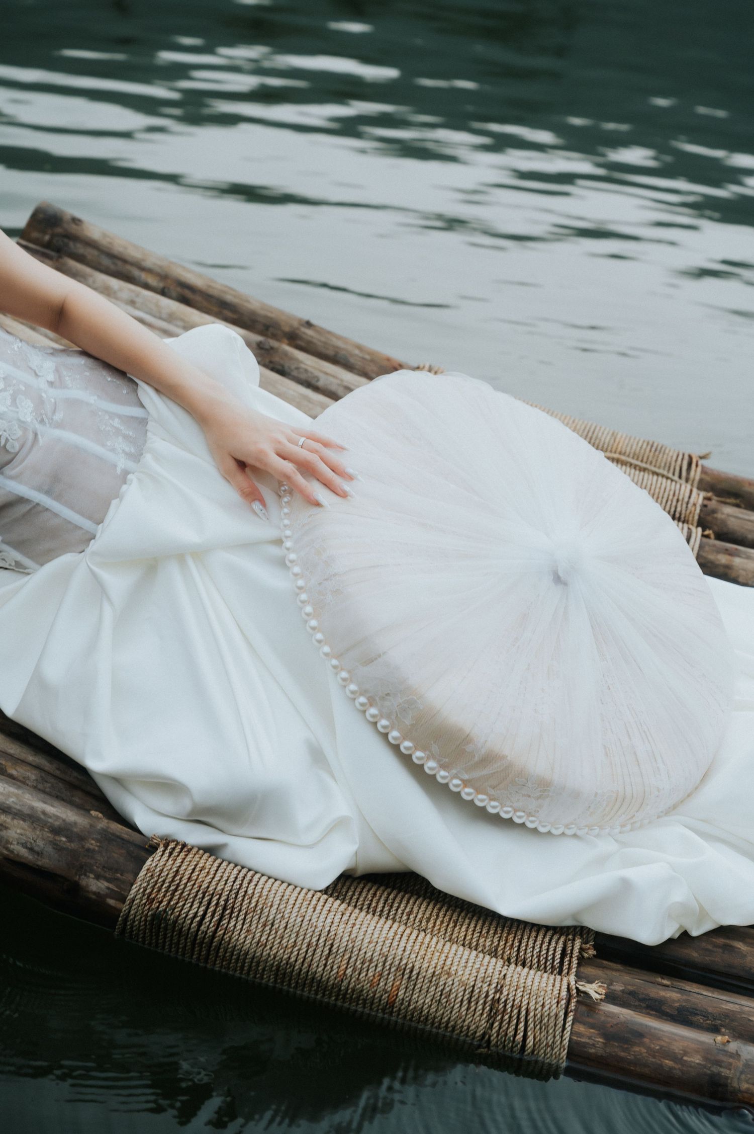 A bride in a white gown reclines on a bamboo raft over water, holding a pearl-trimmed hat.