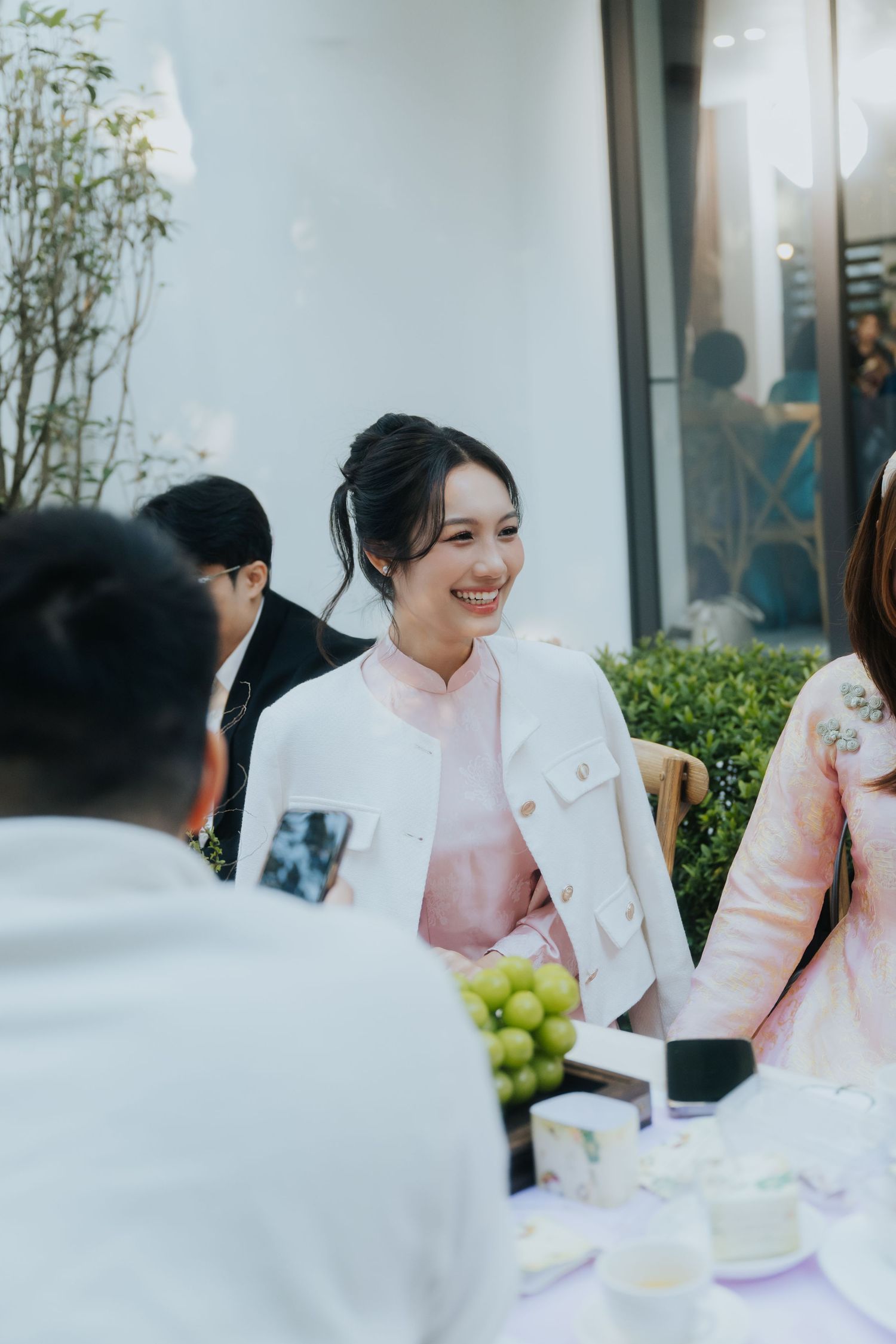 Smiling Asian woman in white jacket and pink dress at an outdoor social gathering with guests around a table.