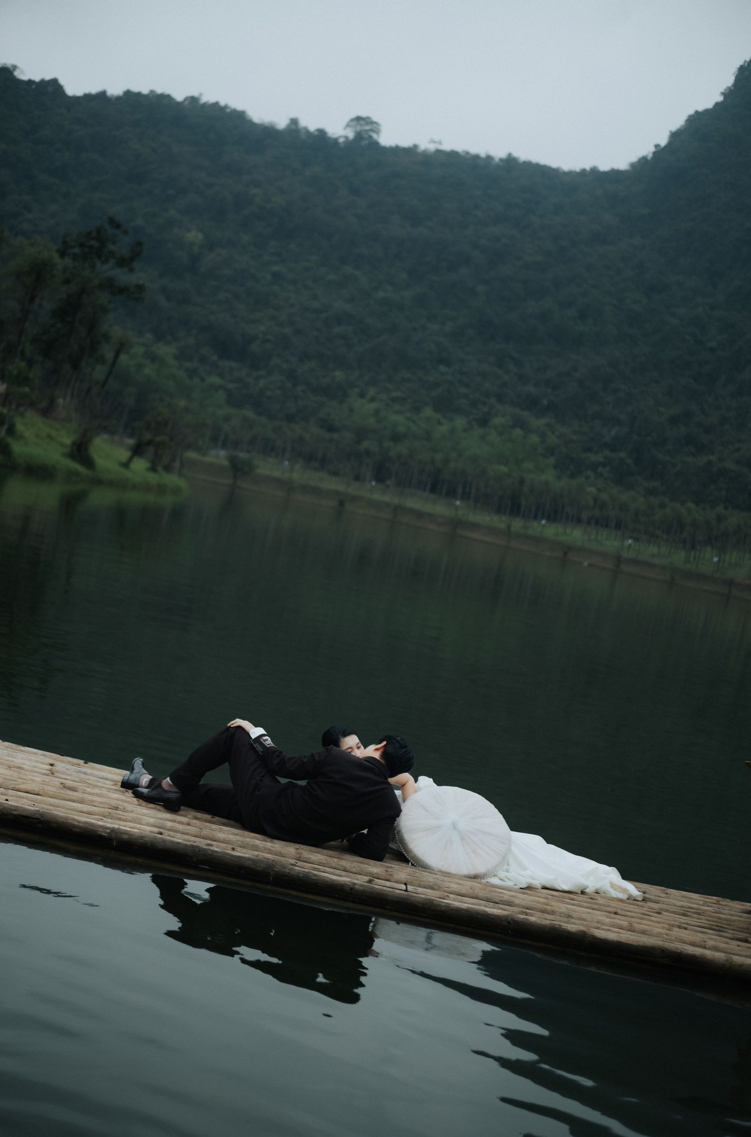 A couple in wedding attire lying on a wooden dock by a serene lake with lush green mountains in the background.