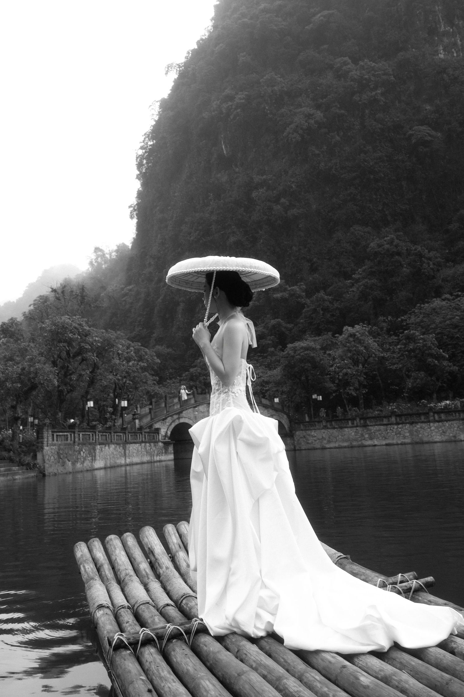 Woman in white wedding gown holding parasol stands on bamboo raft near misty karst mountains in black and white photo.
