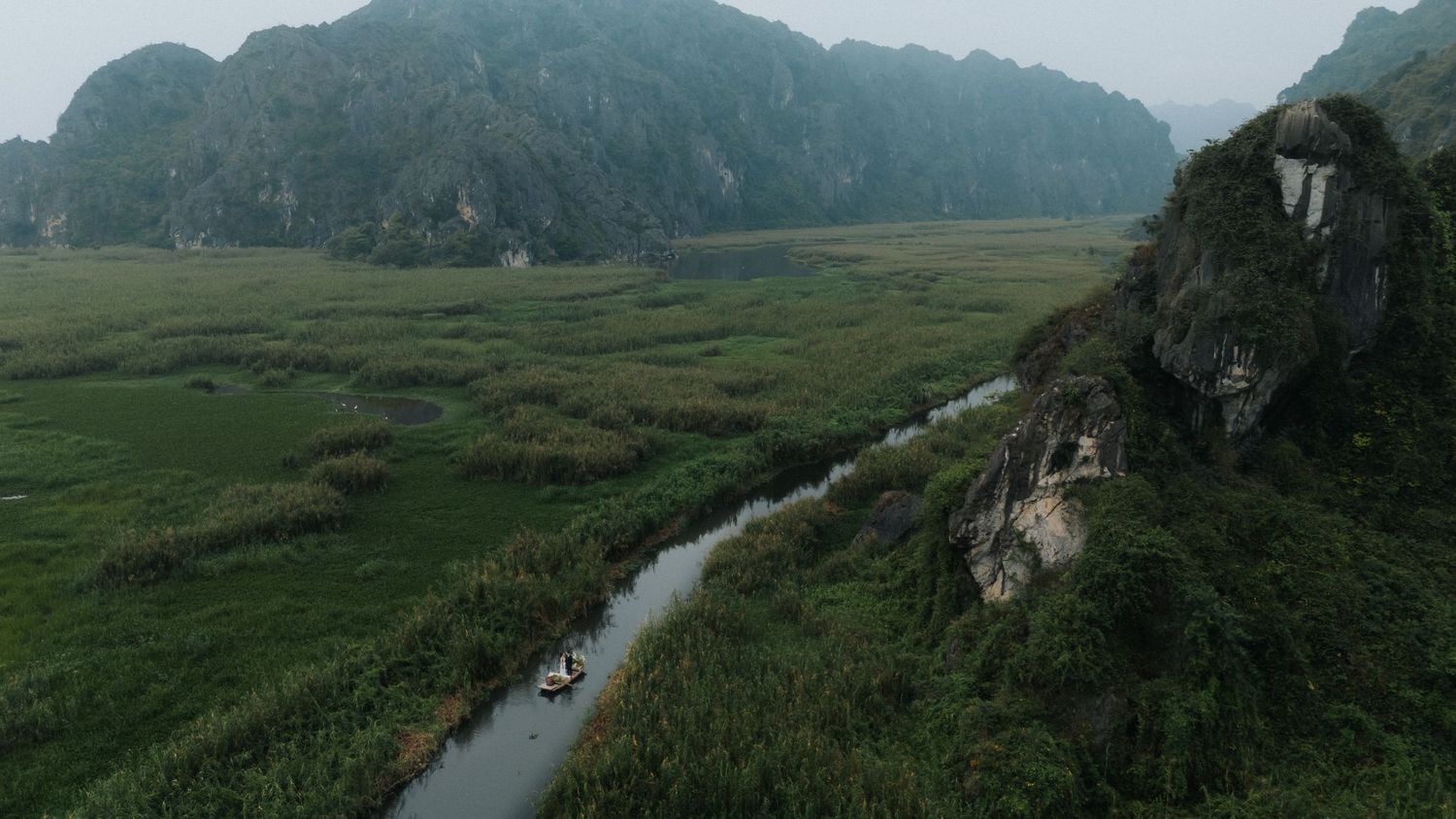 Aerial view of a winding river through lush green valleys with dramatic karst limestone mountains in misty Vietnam.