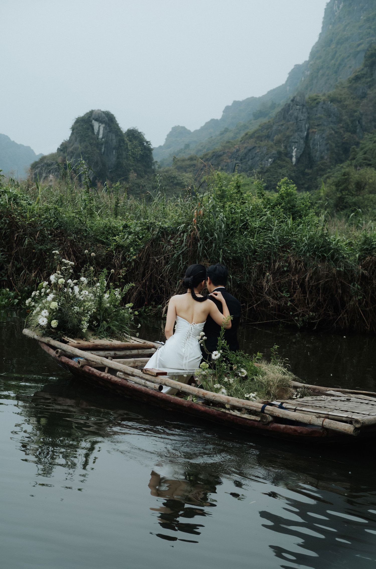 Couple sharing a romantic kiss on a rustic wooden boat adorned with white flowers, surrounded by misty karst mountains.