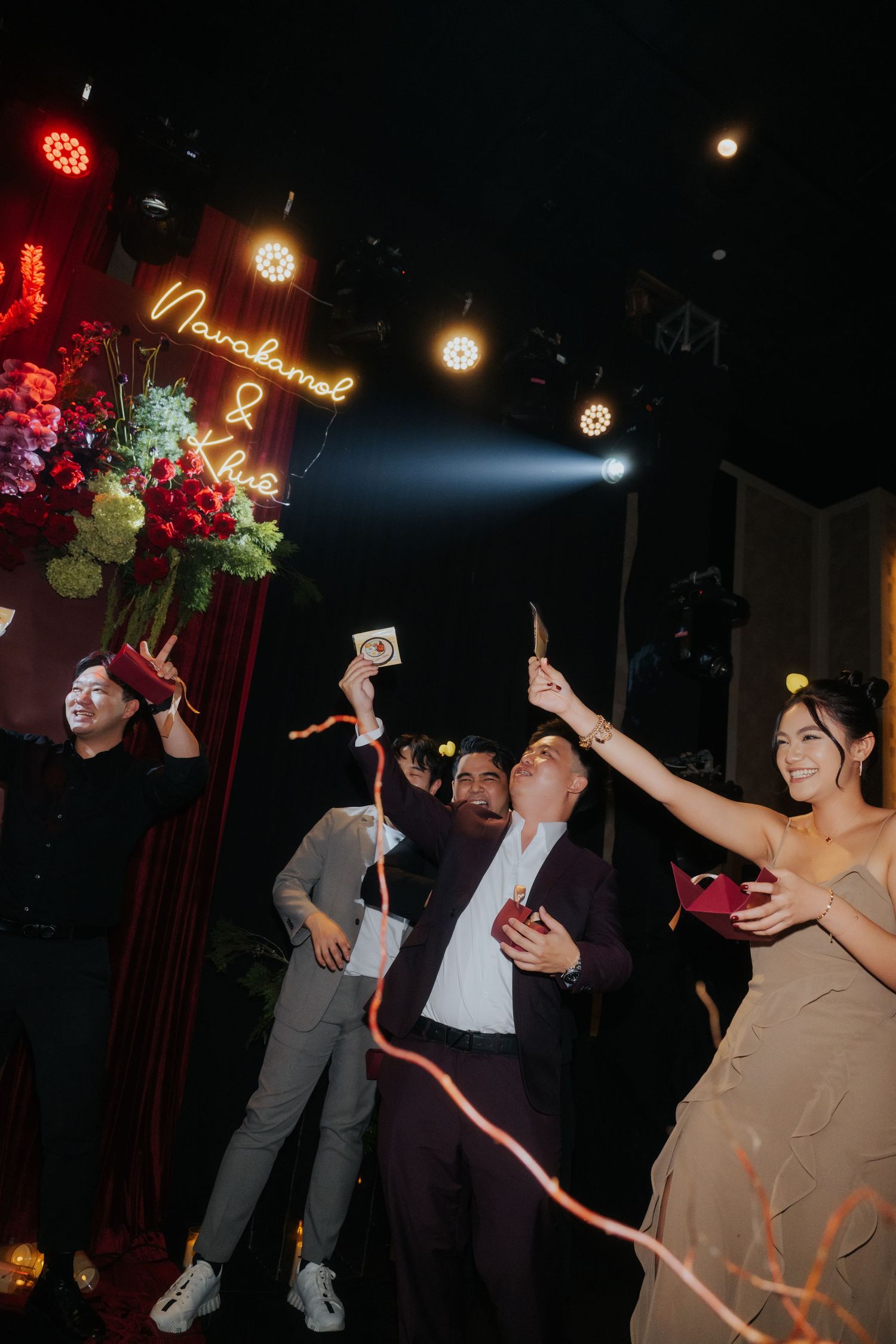 Guests celebrate at a wedding reception under neon signs and festive lights, holding sparklers and cameras.