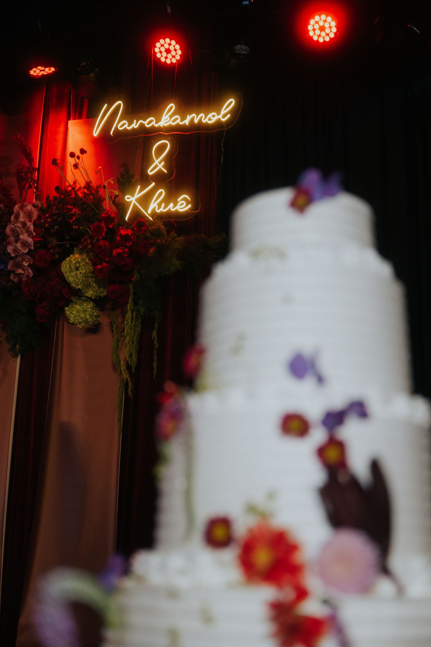 Multi-tiered white wedding cake with floral decorations beside a glowing neon sign reading Narakamol and Khue.