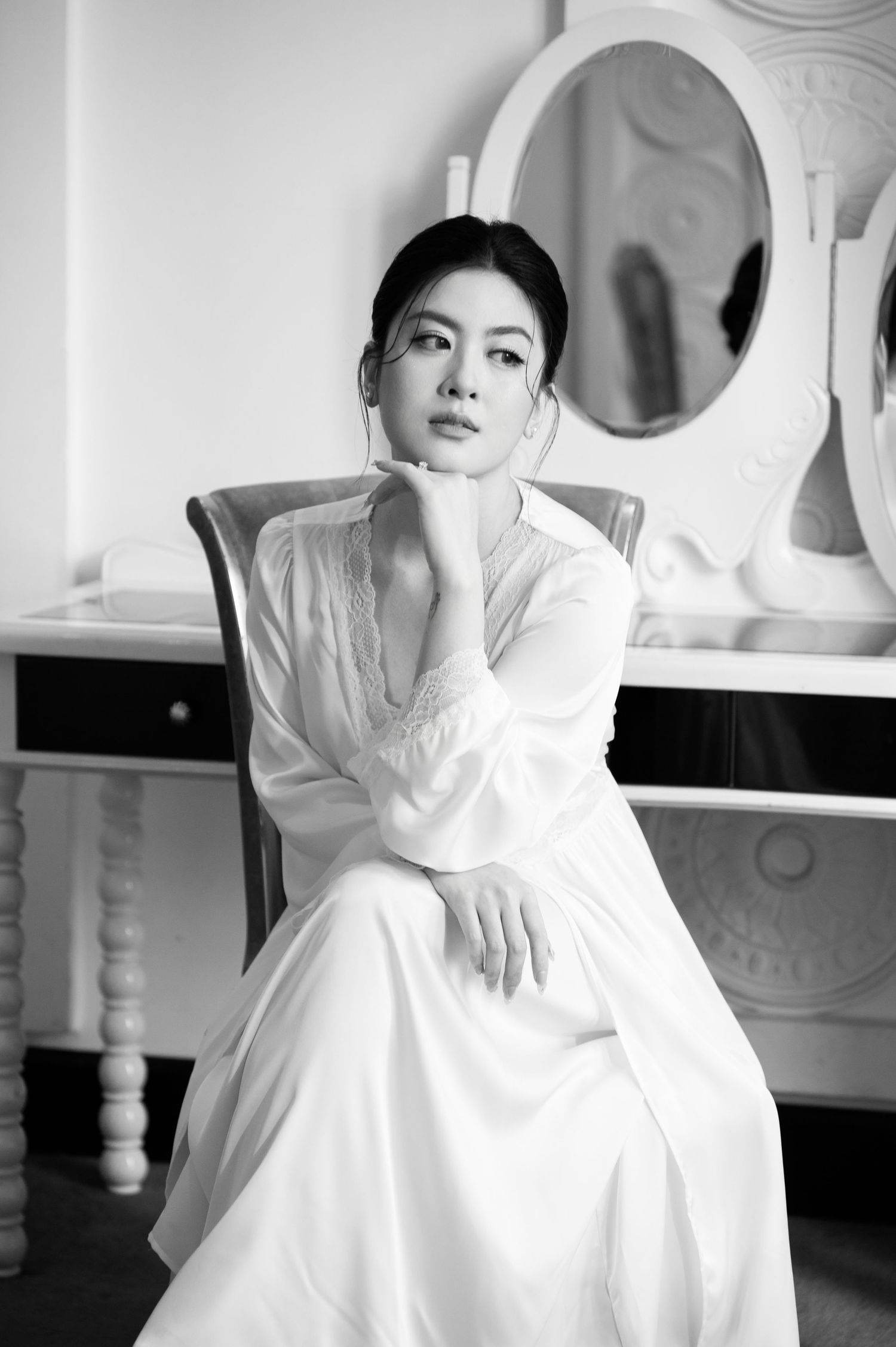 Black and white portrait of an elegant woman in a white dress seated gracefully near a vanity mirror.