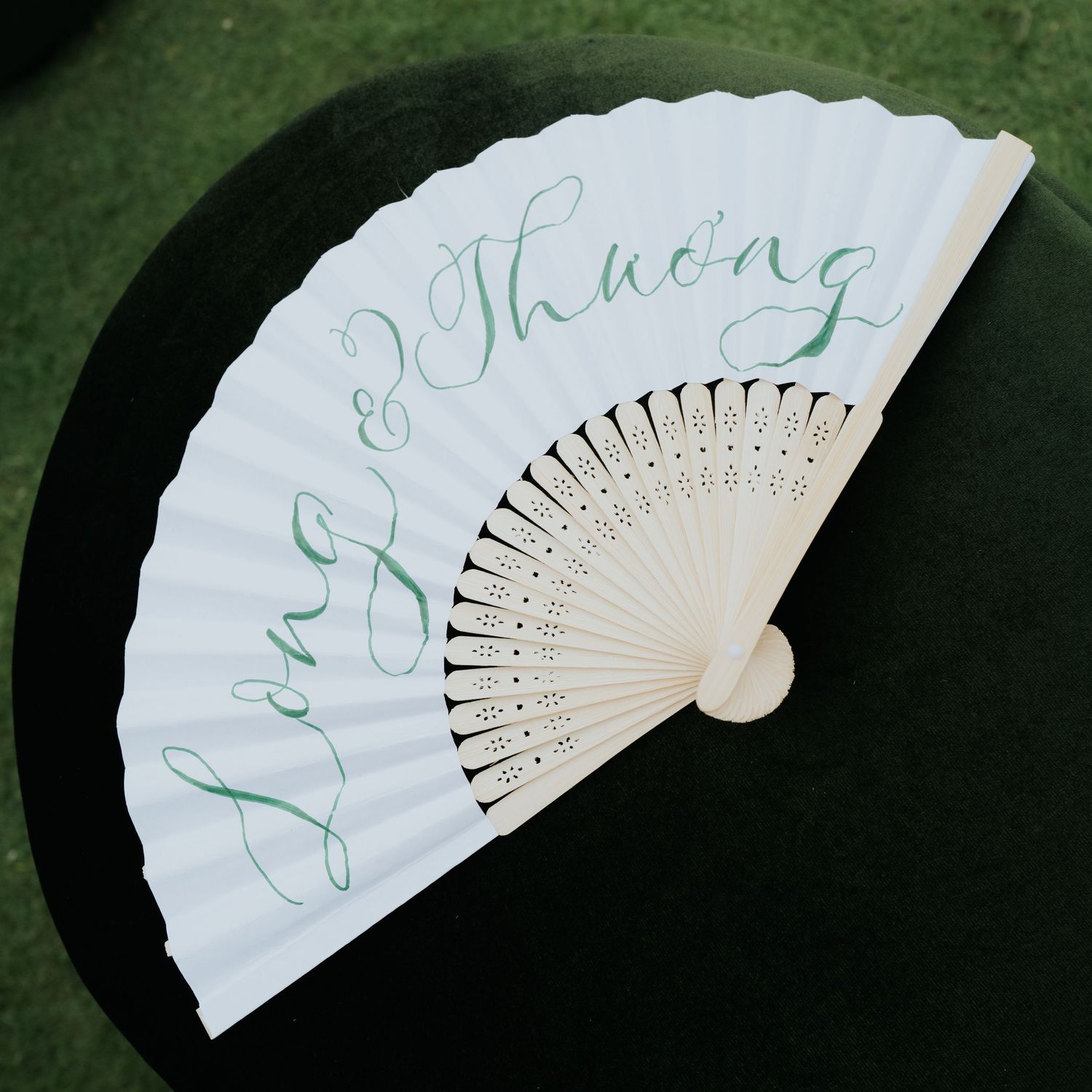 White hand fan with green calligraphy script reading Long & Thuong, displayed open on dark green velvet surface.