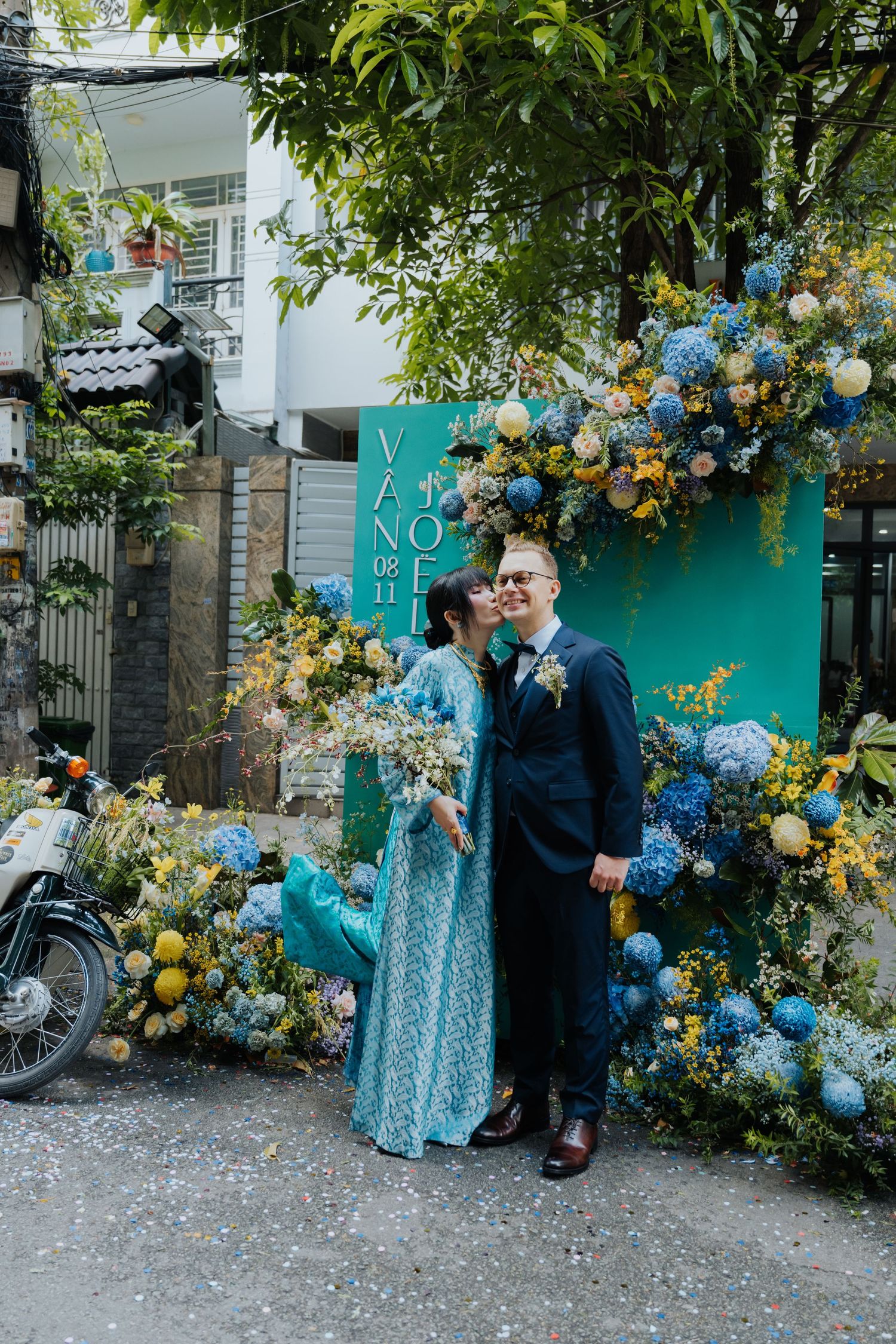 Couple kissing in front of teal floral wedding backdrop with blue and yellow flowers outdoors.