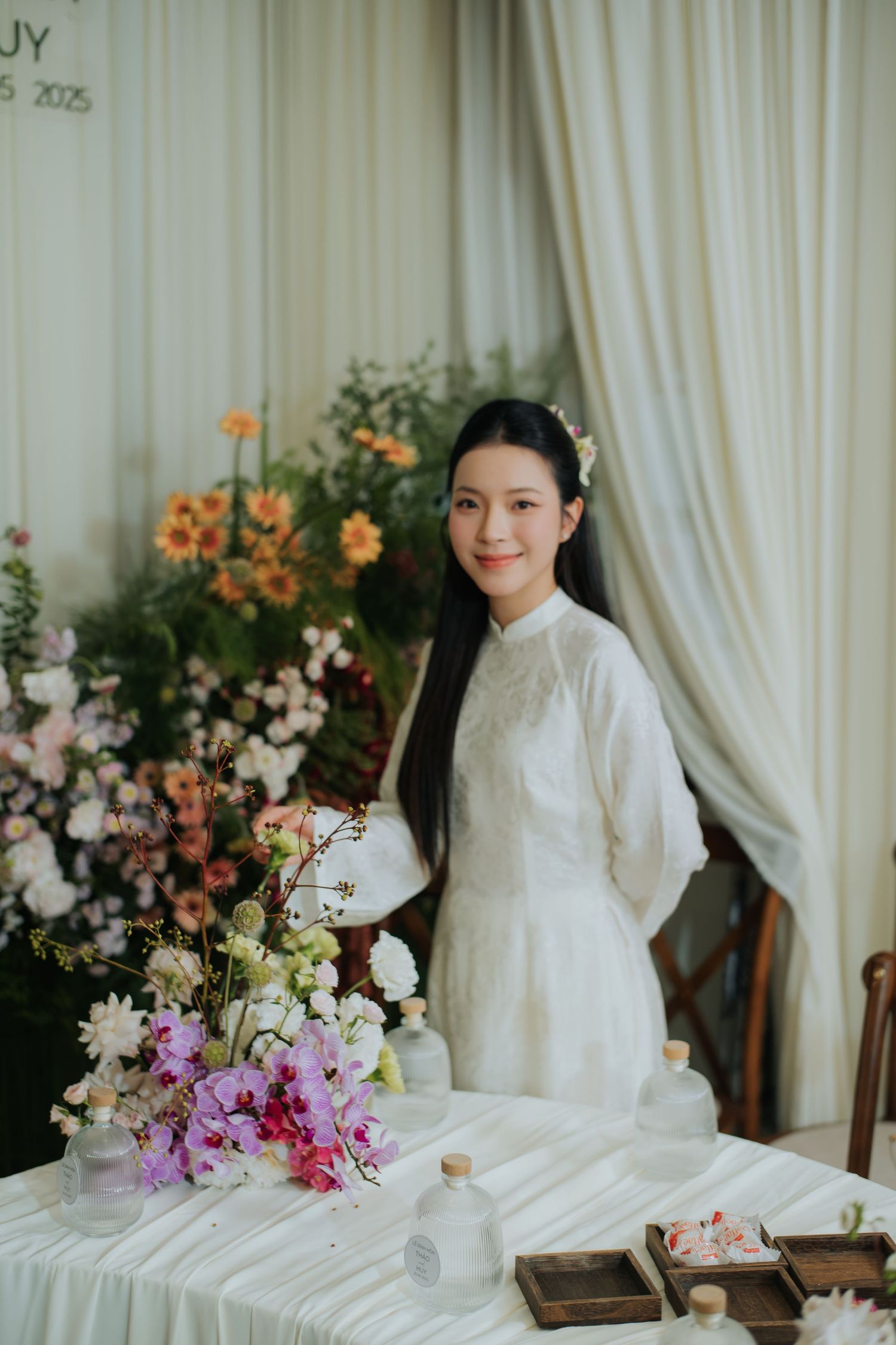 A young woman in a white ao dai dress smiles beside a floral arrangement at an elegant wedding celebration.